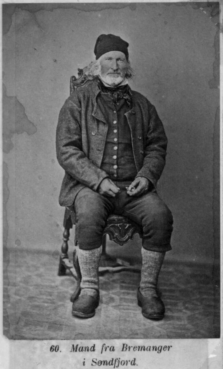 60. Mannsdrakt, Bremanger. Portrett av eldre mann sittende på stol i fotoatelier med nøytral bakgrunn og mønstret gulvteppe. Visittkortformat. Håndkolorert.
Fra M. Selmers (1819-1900) folkedraktserie "Norske Nationaldragter" (nr.60).