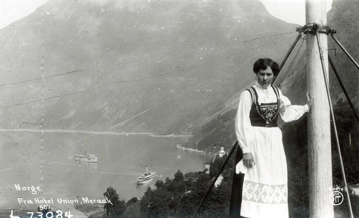 Kvinne står foran fjord med bratte fjell og turistskip.Drakt fra Hardanger.