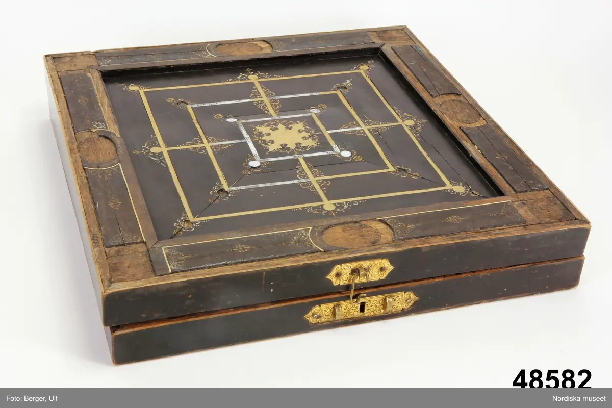 Brädspelslåda för bräde, kvarn och schack, med sju spelbrickor. Svart fanér med inläggningar av pärlemor, elfenben samt graverade förgyllda metallskivor. Blindträ av ek. (Uppgifter från katalogkort). Tillhörande träask för förvaring av spelpjäserna.