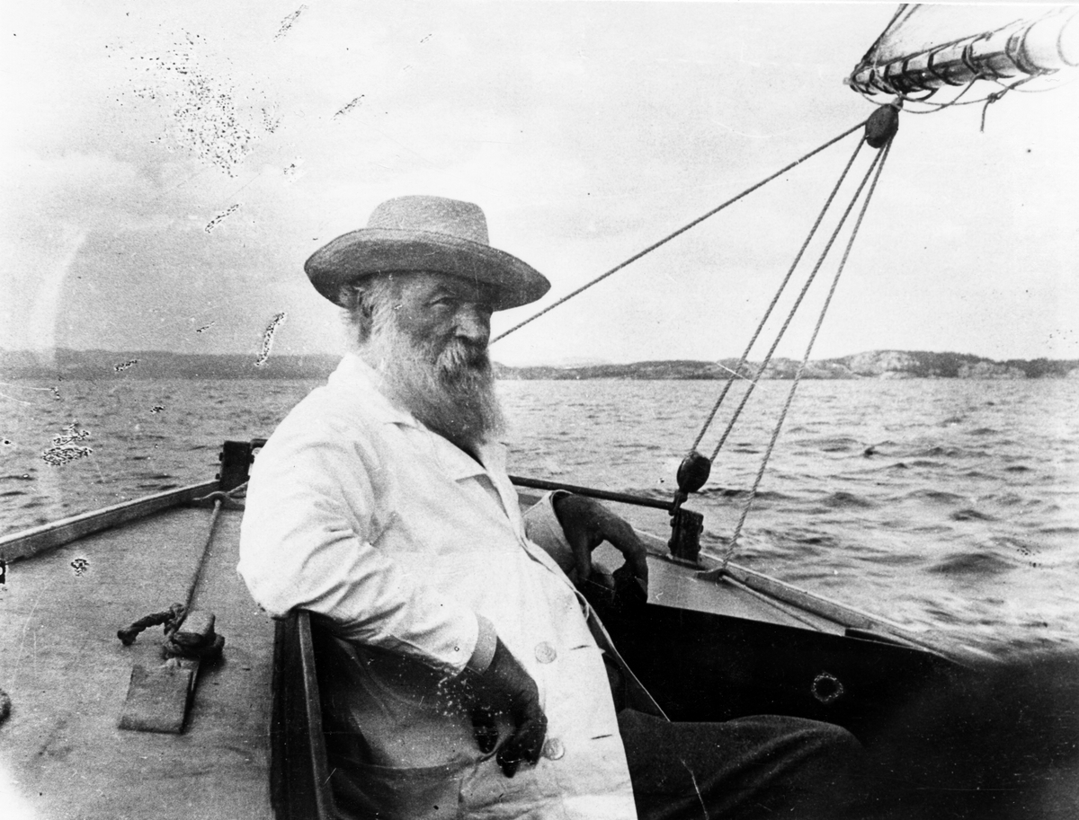 Portrett av båtbygger Colin Archer på lystseilas i Larviksfjorden. Han sitter og holder i roret i en seilbåt, iført en lys jakke og hatt.