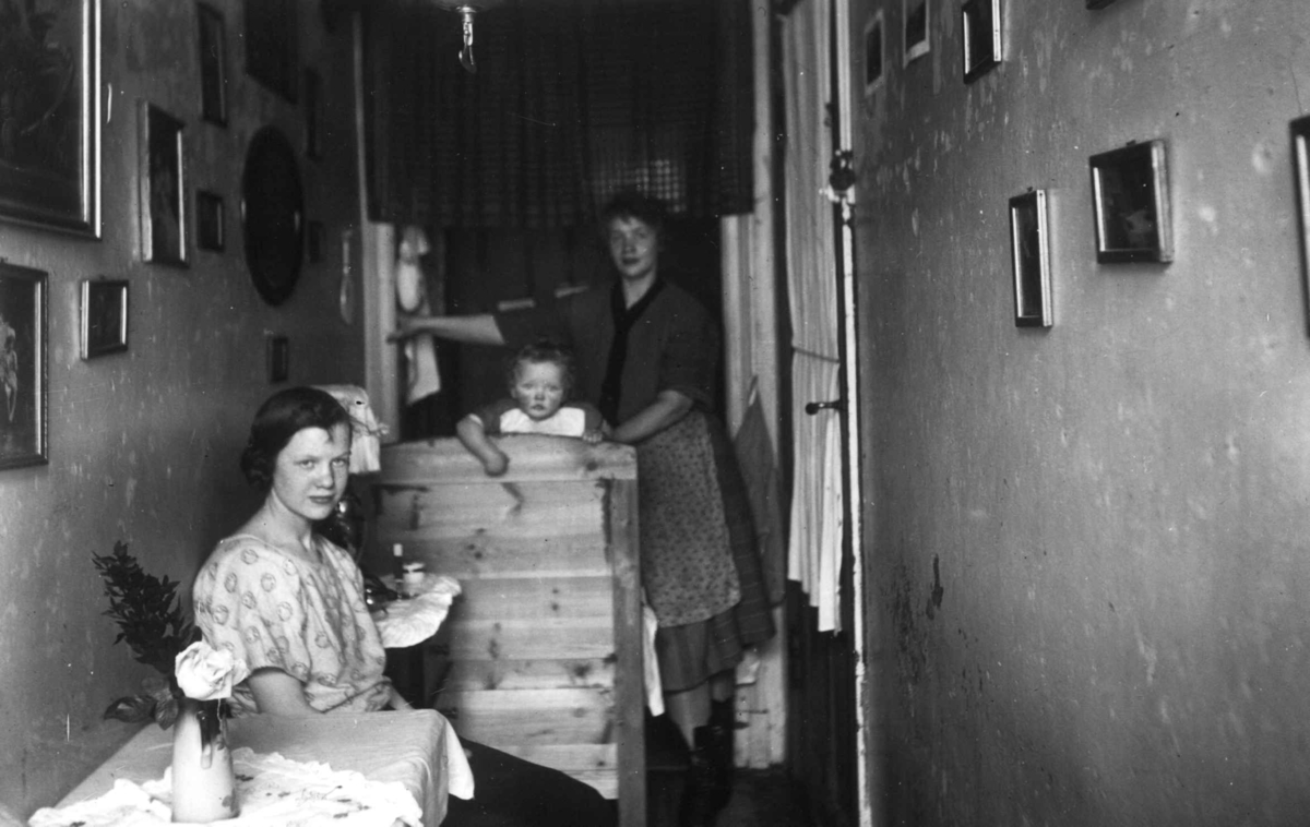 Interiør, bebodd gang, møblert med møbler og seng. Fra Det Rivertzske kompleks, Sagene. Mor med barn poserer.
Fra boliginspektør Nanna Brochs boligundersøkelser i Oslo 1920-årene.