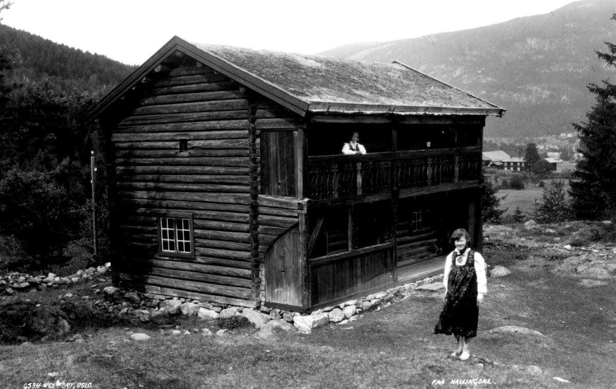 Kvinnedrakt, Nesbyen, Hallingdal. En kvinne står foran et tømmerhus. En annen står i svalgangen i husets 2. etasje. Bebyggelse og fjell ses i bakgrunnen.