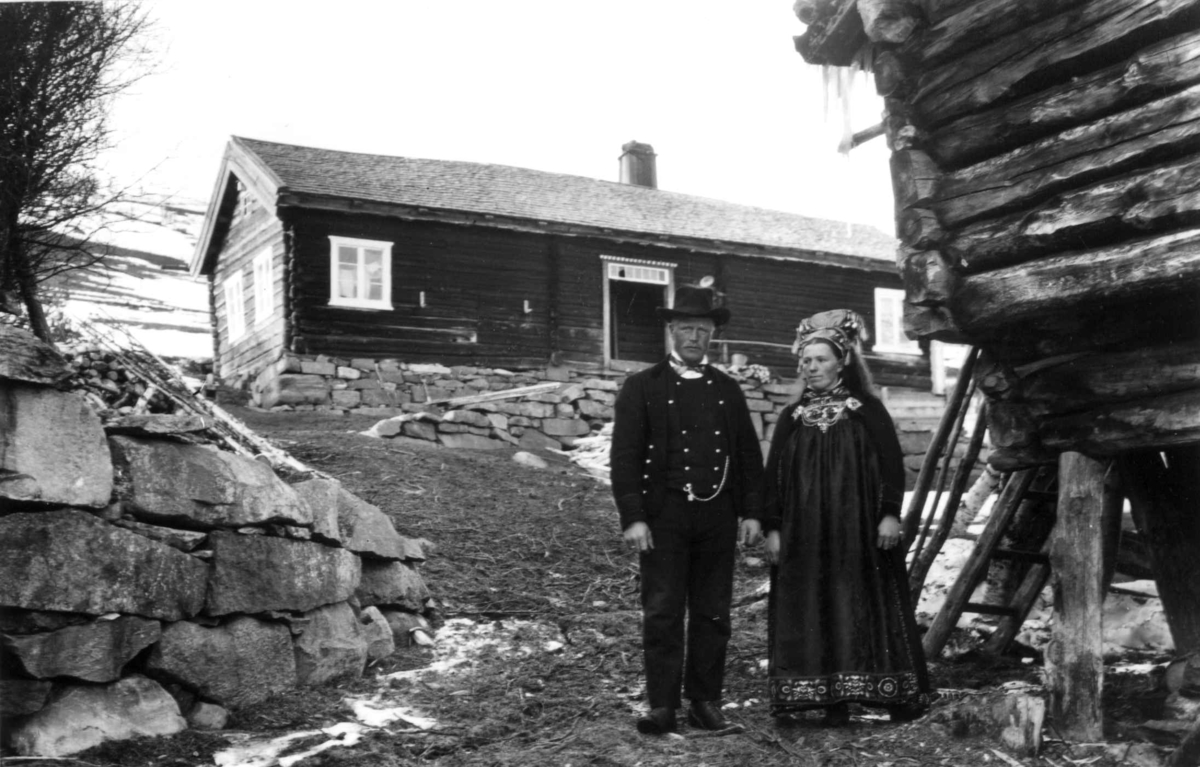 Manns- og kvinnedrakt. Mann og kvinne står på gardstunet.  Litt snø er igjen etter vinteren. Tømmerhus ses i bakgrunnen.
