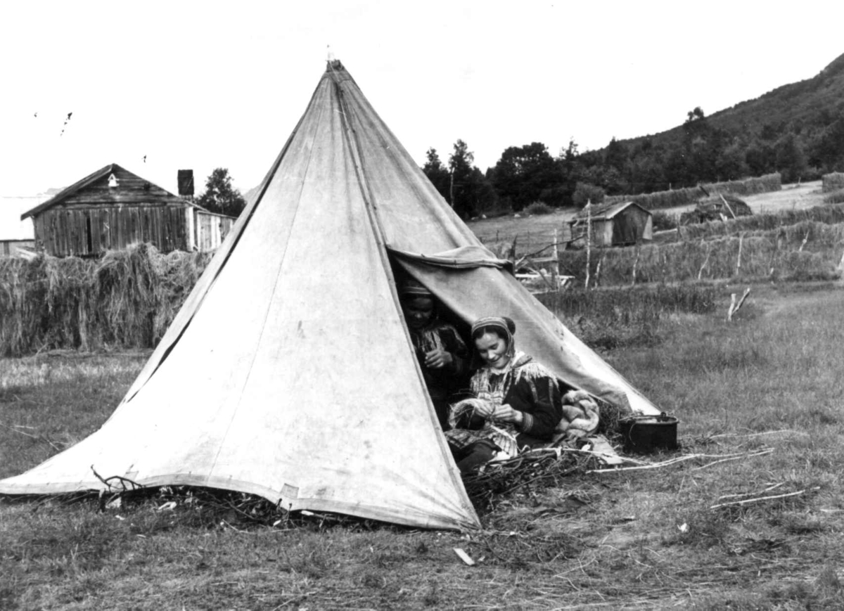 To kvinner i åpningen til et sametelt, Hesjer og bygninger i bagrunnen. Olderdalen, Kåfjord, Troms 1947.