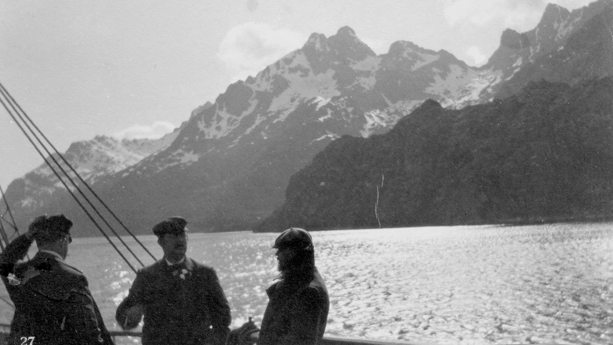 Tre menn på dekket av båt. Fjord og fjell med snø. Vestsiden av Raftsundet.