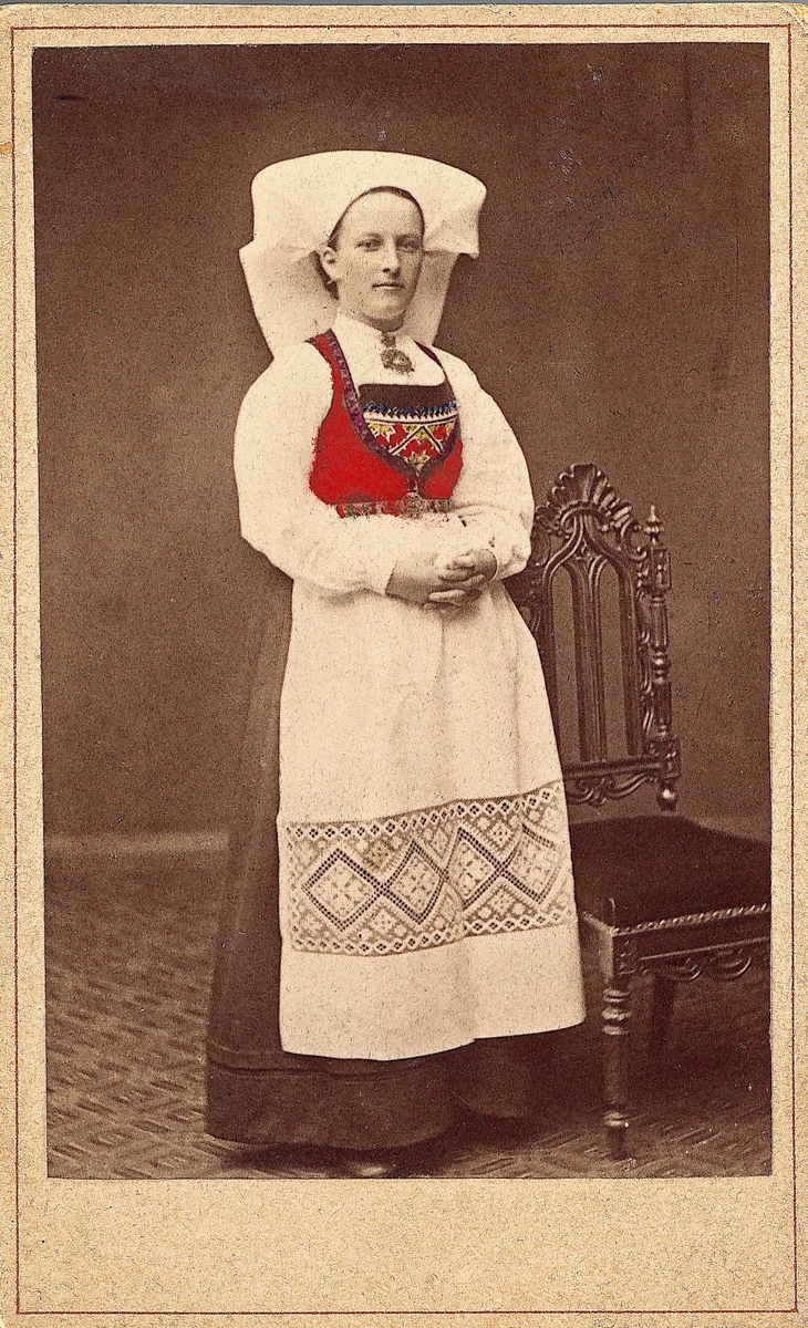 U.nr. Konedrakt fra Hardanger. Portrett av kvinne stående ved høyrygget stol ant. i fotoatelier med nøytral bakgrunn og mønstret gulvteppe. 
Fra serien "Norske Nationaldragter", visittkortformat, håndkolorert. Serien er tatt av fotograf Marcus Selmer (1819-1900), Bergen.