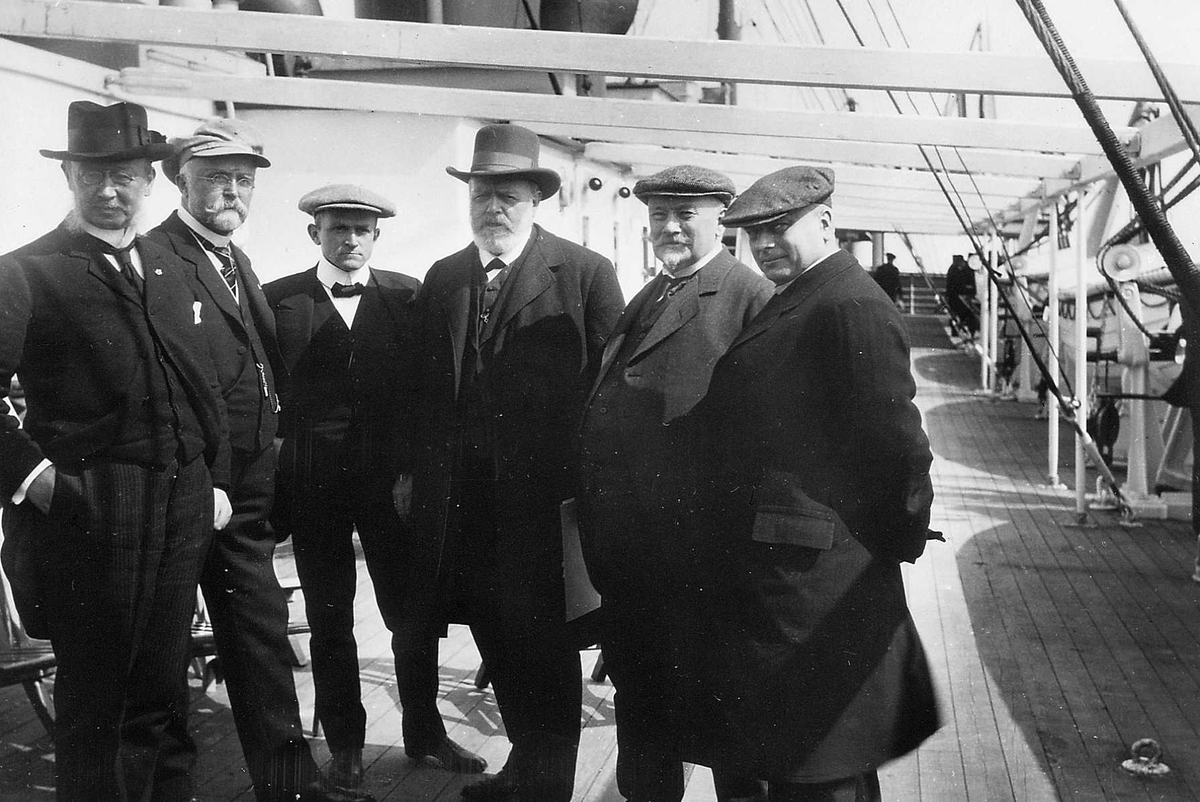 "6 Arbeidsministere Lehmkuhl, Brænne, Darre-Jensen, Løvland, Ihlen, Urbye" - portrett av 6  menn i dress eller frakk og med ulike hodeplagg på dekk. Motivserie fra Den Norske Amerikalinjes (NAL) D/S "Kristianiafjord"s første tur Kristiania - Bergen 1913, tatt av fotograf  Anders Beer Wilse, samlet i et album utgitt på  kunstforlaget Mittet & Co.