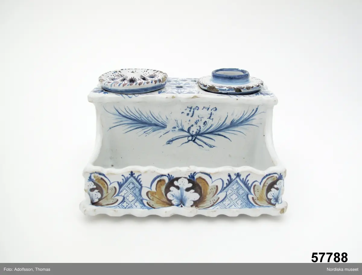 Katalogkort:
"Skrivtyg, fajans. Rektangulärt, framtill rum för pennor, upptill bläckhorn och sanddosa; dekor. med blommor och rutmönster i blått, brunt och gult, på framsidan två palmkvistar i blått, varöver H.H. 
Sign. i blått: Stockholm 2/8 1749 Hindrich Höök.
/Upmark 1904"

Skrivställ av fajans med dekor i blått och manganviolett, Rörstrand 1749.
Skrivstället med runda dosor för bläck och strösand är målat av Henrik Höök som signerat det med hela sitt namn under botten, oftast står bara målarens initialer utsatta. Han var då lärgosse vid fabriken och blev målargesäll 1754. 
Märkning: "Stockholm 2/8 1749" samt "Hindrich Höök".
Byggmäst E A Bomans samling nr  75.
/Annika Tegnér 2006