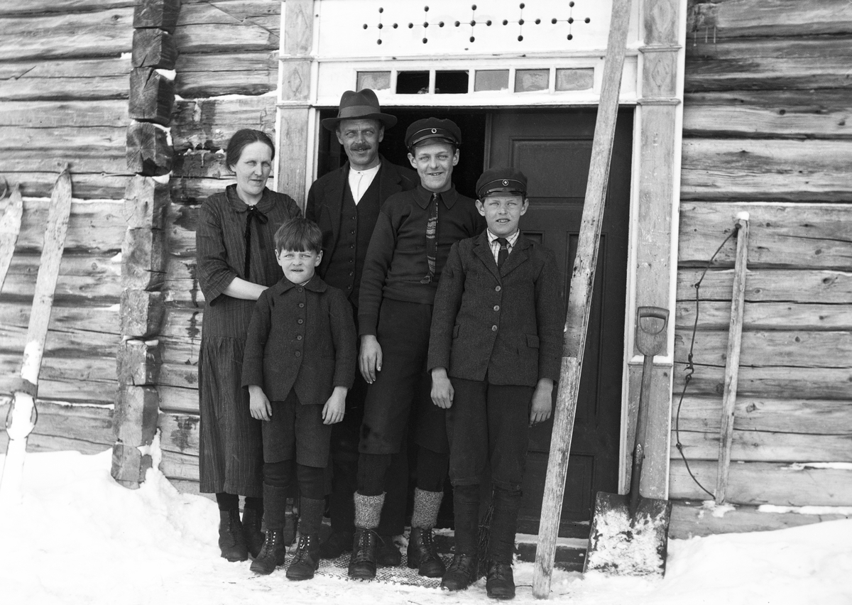 En familie med mor, far og tre gutter står døråpningen, ski står opp mot veggen på huset.