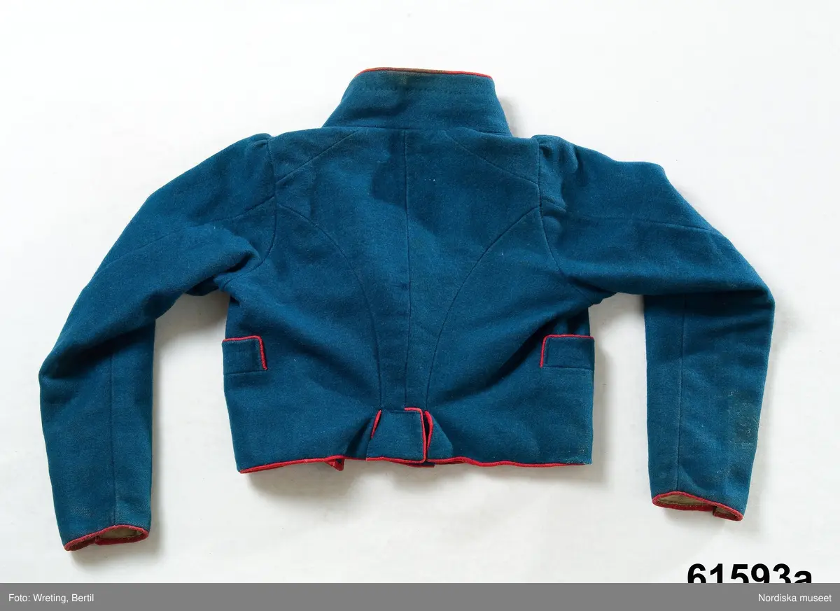 Huvudliggaren:
"Mansdräkt från Forsa sn."

A. Jacka längd
B. Knäbyxor längd 64,5 cm, benlängd 42 cm, luckbredd 48 cm, luckhöjd 11 cm
C. Väst längd 53 cm, kraghöjd 4,5 cm
D. Väst längd fram 44 cm, kraghöjd 7,5 cm
E. Mössa höjd 28 cm, omkrets 56 cm, uppslag 9-10 cm

A. Jacka, av mellanblå vadmal, 2 framstycken, 2 avsmalnande ryggstycken som slutar iskörtflikar. Isydd ärm formad med 2 sömmar och rynkor på ärmkullen, ståndkrage, runt alla kanter röd passepoal av ylletyg, knapphål sydda med röd lintråd på båda framstyckena, dubbelknäppt med 12 knappar klädda med rött tyg, helfodrad med oblekt linnelärft, 2 iskurna fickor med rödkantade ficklister. Malhål i kanterna.

B. Knäbyxor av svart vadmal, bred lucka med 4 knapphål Midjegjorden knäpps fram med 2 par knappar och knapphål, sprund bak med 2 par snörhål och sämskskinnsrem för hopsnörning.
6 mässingsknappar. Vid knät sprund knäppt med 5 ornerade mindre mässingsknappar. Knälinning knäppt med tränsat knapphål samt svartbrun knapp. Alla knappar fästade med sämskskinnsremsor. Helfodrade med halvblekt linnelärft.

C. Väst av randigt halvylle i korskypert med smala bomulls- och silkeränder. 2 framstycken, 2 bakstycken, krage med slag, dubbelknäppt med 6 par toppiga mässingsknappar med grönt mönster, 2 par knappar vid krage och slag, 2 stolpfickor, foder av grov oblekt linnelärft.
1 knapp saknas.

D. Väst av vit randad bomullspiké, ryggstycke av vit linnelärft, som även är foder i framstyckena. Dubbelknäppt med 9 par små överklädda knappar i samma tyg, två stolpfickor. Hög ståndkrage.

E. Toppmössa stickad av rött ullgarn i slätstickning och filtad.Uppslaget vidgat, mössan dubbel, i toppen en silketofs i blågrön färg. Sannolikt maskinstickad och färdigköpt.

Berit Eldvik 2004
