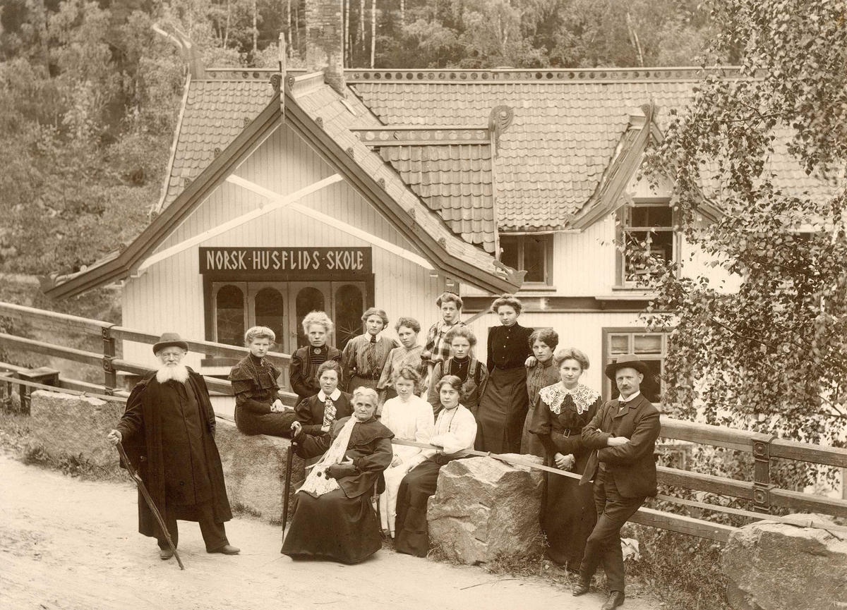 Gruppeportrett. Menn og kvinner står utenfor norsk husflids skole.