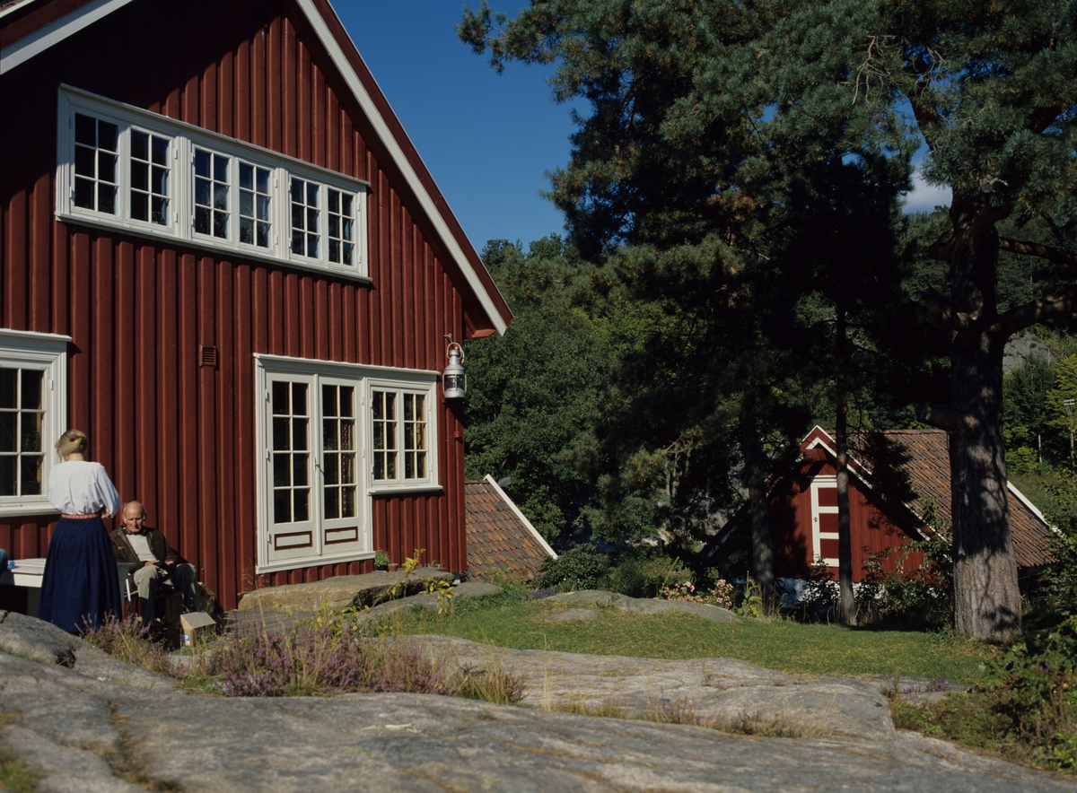 To personer sitter på en terrasse ved endeveggen på bolig og et brønnhus i bakgrunn ved Normannsvik utenfor Tvedestrand. Illustrasjonsbilde fra Bonytt 1986.