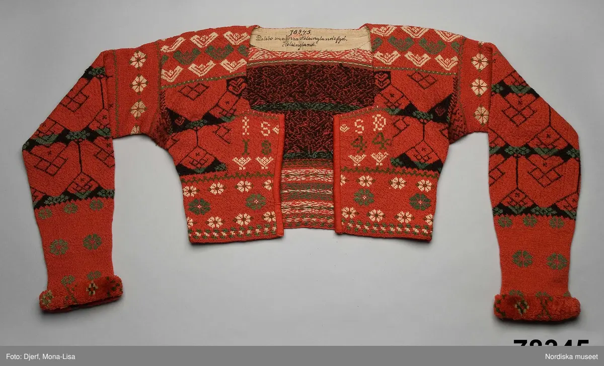 Kvinnotröja, midjekort, slätstickad i hårt tvinnat tunt ullgarn, helt mönstrad i rött som dominant färg, samt svart, grönt och vitt. Det vita garnet är bomullsgarn, vilket föredrogs för att det gav en renare vit färg än ullgarn. Mönstret är specifikt för socknarna Delsbo och Bjuråker. Stickningen utförd så att trådarna binder hela vägen på baksidan. Bålen rundstickad och sedan uppklippt mitt fram, där öppningen kantats med rött kläde. Fyrkantig halsringning infodrad med oblekt linnelärft. Knäppt med 5 par hakar och hyskor av mässing. Ärmarna stickade för sig och vidsydda mot bålen. Ärmarna avsmalnar mot handleden där de kantats med ett flossabräm av  ullgarn i rött med mönster i grönt och vitt. Brämet vävt med botten av lingarn och inknutna nockor som sedan klippts kort och jämnt.
På bröstet instickad märkning I S S D  1 8 4 4 = jungfru Sigrid Svensdotter.
Dessa tröjor blev moderna kring 1830-40 tal i Delsbo och användes som söndagseftermiddagsdräkt framförallt av de unga som då samlades till dans eller andra nöjen. Männens tröjor var av samma slag men var inte öppna fram.
/Berit Eldvik 2007-07-03