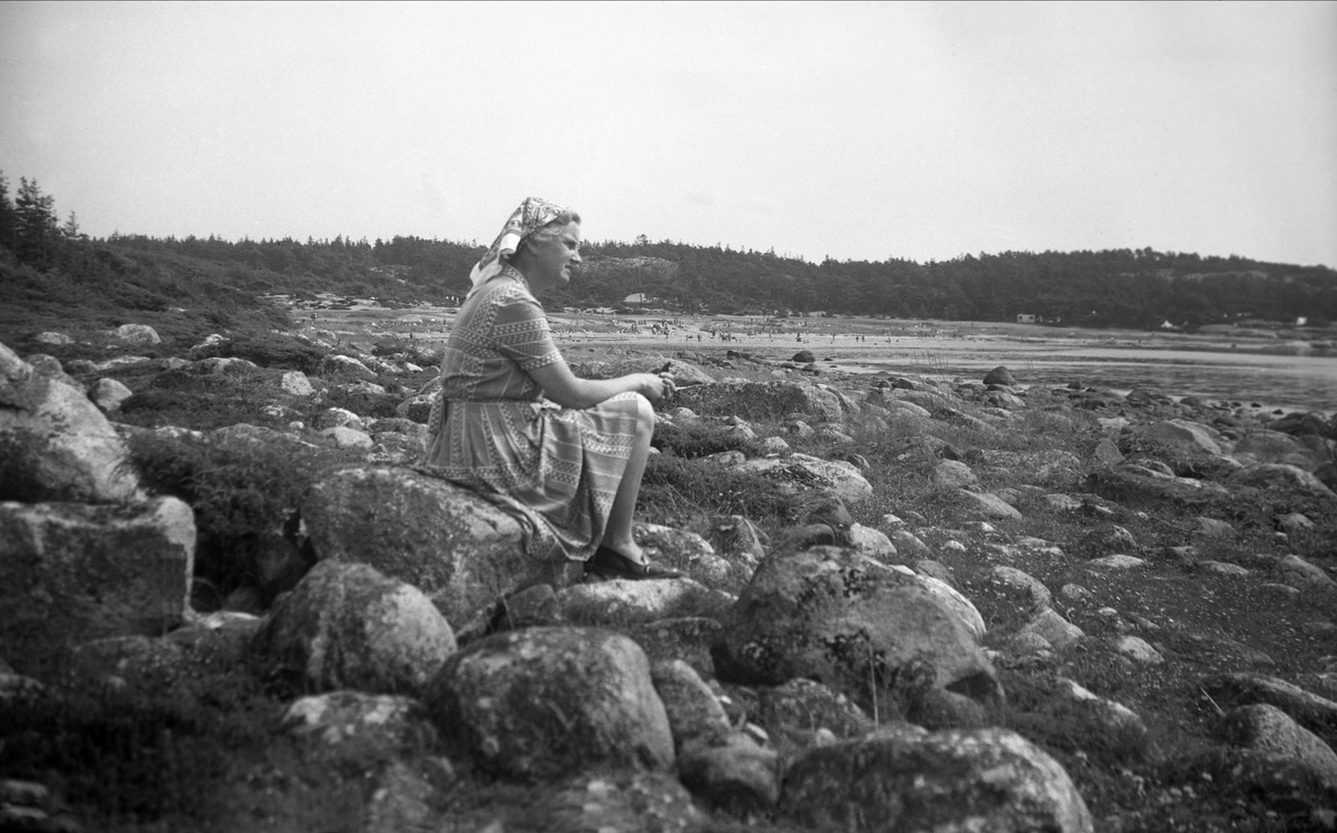 Dordi Arentz sitter ved vannkanten på Hvaler sommeren 1950. I bakgrunnen badeliv på en av øyas strender.