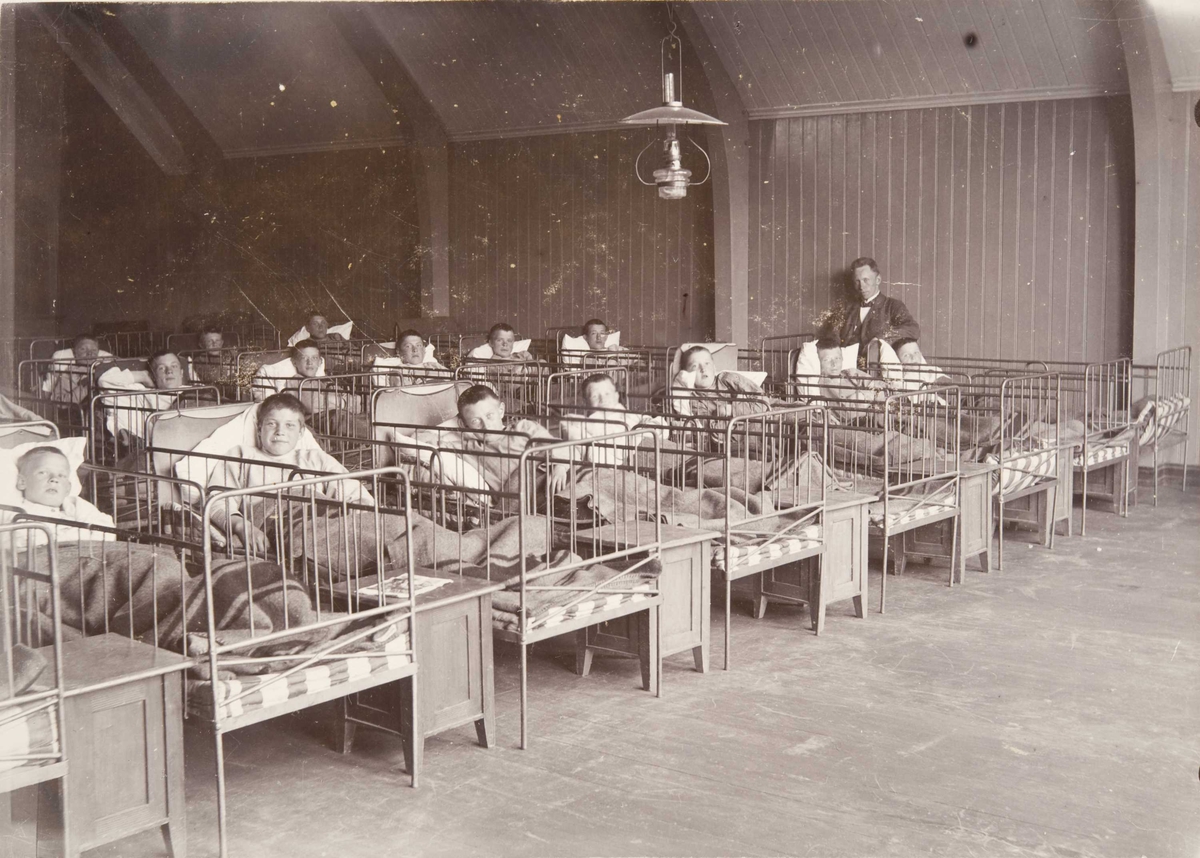 Sovesalen på Bastøy skolehjem, interiør, senger, Bastøya, ca. 1903. Elever og en ansatt.