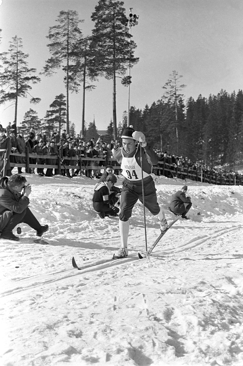 Skiløper med startnummer 94 i Holmenkollen. Publikum langs løypa.  Pressefotografer på sidelinjen. Holmenkollrennene 1963.