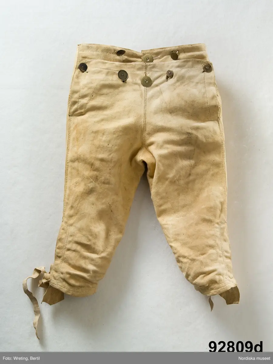 Mansdräkt i flera delar a-d.

a. Rock av svart vadmal
b. Röd väst
c:1-2 Vita strumpor
d. Knäbyxor av sämskskinn
Katalogkort finns.

a. Långrock av svart vadmal. Framstyckena/sidstyckena helskurna, ryggstycket smalt och kort,  avkuret i midjan, rikt veckat bakskört försett upptill med två ulltofsar och med pikerade klaffar en vid var  sida om skörtet. Svängda ärmar med 2 sömmar och blindslag med sprund oknäppt med 2 hakar och hyskor. Ståndkrage. Skoning av hemvävda ylleband i flera färger längs livets insidor, knäppning med hakar och hyskor.
"Dylika rockar tillhörde högtidsdräkten, vid mindre fester och till kyrkan brukades grå rockar af liknande snitt, ett snitt som gaf anledning till benämningen ´fiolen´för nysockenströjans ryggtafla ".
b. Väst av röd vadmal med ryggstycke av grovt oblekt linne i spetskypert. Hög ståndkrage och utvikta slag, med en uddklippt vadmalsremsa i kanten, dubbelknäppning med  vitmetallknappar, fästa på insidan med skinnremsa, knapphål tränsade med grönt ullgarn. 2 sidfickor med list. Foder av oblekt linnelärft, på kragens insida en bit av ryggtyget mitt bak.
c:1-2 . Strumpor slätstickade i tvåändstickning av fint vitt 1-trådigt ullgarn. Uppläggningsvarvet med rött och vitt ullgarn,  smal mönsterstickad överkant. Häl och tå av annat naturgrått garn. Mitt bak en sömmarkering med en avig maska. Vid anklarna broderade rombiska figurer i genombrutet mönster, kallas "löver" i rött ullgarn.
d. Knäbyxor av gult sämskskinn, med bred lucka med 5 knapphål, midjegjord knäppt med 2 knappar. Knapparna är av flera olika sorter av mässing, en av hängselknapparna av horn, 2 bak saknas. Midjegjorden fodrad med linnelärft. Knäsprund knäpps med 4 hakar och hyskor. Flikar vid sprunden uddklippta.
/Berit Eldvik 2010-01-08
