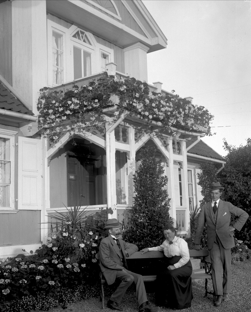 Gruppe, to menn og en kvinne ved en benk foran inngangen på en villa. Blomster rundt og på verandaen.