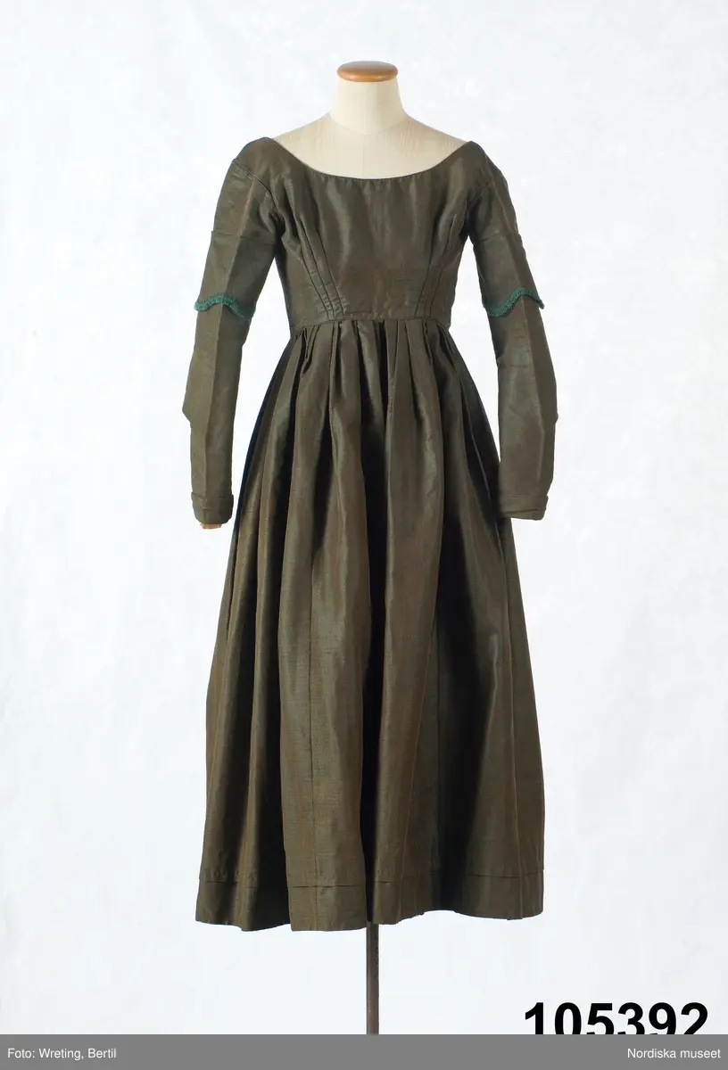 Hel klänning i 1830-talets modesnitt, av fint halvylletyg i kypert med varp av ljusbrunt bomullsgarn och inslag av grönt tunt glansigt redgarn som ger en grönskimrande yta. Livet går ned nästan till midjehöjd, figursytt med 2 framstycken med bakåtflyttade sidsömmar, ett ryggstycke med 6 inprovningar fördelade i två grupper. Vid båtringad halsringning, knäppning med tätt sittande hakar och hyskor. Isydda långa ärmar svängda med 2 sömmar  med 3 veck vid armbågen. Ärmsprund knäppt med hyska och hake samt 4 cm uppslag med rundade hörn. Upptill på ärmen besättning av tyget med stora rundade uddar kantade med klargrön bomullsfrans.  Passepoaler av tyget i alla sömmar. Livet fodrat med skotskrutig bomullslärft i rött, grönt, gult och vitt och i ärmarna ett smalrandigt bomullstyg i blått och rosa. Kjolen sydd av  5 våder, lagda veck runtom i midjan. sprund mitt fram. Uppsytt veck  en decimeter från fållen. Skoning med brun bomullslärft. Handsydd.
Har burits 1838 som ungmorsklänning, alltså av bruden dagen efter bröllopet, av Maria Margareta Mårtensdotter från Kysings, Vall sn på Gotland. Möjligen tillsammans med sidenhalsklädet 105385.
Anm. I  gott skick.
OBS Klänningen sitter bakochfram på fotona, knäppningen är fram.
/Berit Eldvik 2011-06-13