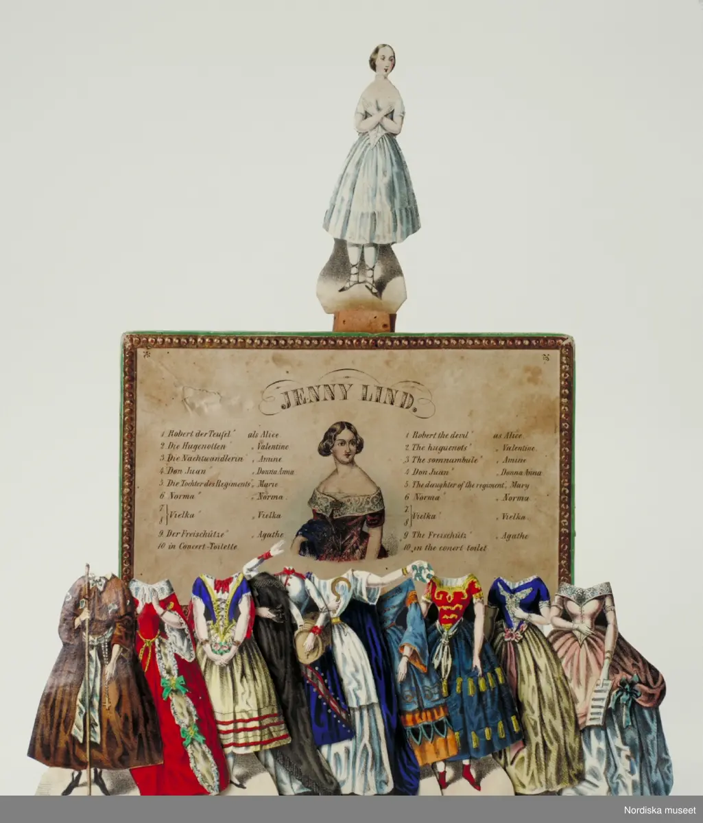 Inventering Sesam 1996-1999:
H 17, B 10,8 cm (kartongen)
Pappersdocka av kartong fodrad med grönt papper, tryck och handkolorerad, föreställande den svenska sångerskan Jenny Lind (1820-1887), som slog igenom stort på 1840-tal. Dockan ligger i sin originalkartong tillsammans med en fotställning av kork och tio kolorerade dräkter.
Dockan (H 10 cm) har gul-brunt uppsatt hår och är klädd i en underkjol och ett snörliv i vitt, vita strumpor och svarta skor snörda upp på vaderna. Hon håller armarna i kors över bröstet. Originalkartongen består av en underdel och ett separat lock, materialet är kartong, grön undertill, rosa på sidorna och ovanpå en guldpappersram med en kolorerad bild av Jenny Lind i röd klänning med spetskrage och hennes namn skrivet som överskrift. På var sida om bilden står skrivet på tyska respektive engelska tio operor och huvudroller som Jenny Lind sjungit och som kläderna i kartongen hör till.
Operor, roller och tillhörande dräkt:
1. Robert the Devil, rollen som Alice, klädd i en brun dräkt, vitt band runt midjan med ett radband och en vandringsstav.
2. The Huguenots, rollen som Valentine, klädd i en röd klänning med vida ärmar och spetskrage.
3. The somnambule, rollen som Amine, klädd i folkdräktslik klädsel med vit blus, blått snörliv och gul kjol med röda ränder.
4. Don Juan, rollen som Donna Anna, klädd i vit klänning och svart mantel.
5. The daughter of the regiment (Regementets dotter), rollen som marketenterskan Mary, klädd i en vit uniformsjacka och en blå kjol, bärande på en öltunna.
6. Norma, rollen som Norma, klädd i en vit klänning och en blå mantel.
7-8. Vielka, rollen som Vielka, klädd i en duvblå och gul klänning med vackra broderier och vida ärmar, samt en dress med rött liv med gulddekoration och en mörkblå kjol.
9. The Freisch¿tz, rollen som Agathe, klädd i en blå överdel, ett rosa band runt midjan och en gul kjol.
10. konsertklänning, blekrosa klänning med spetskrage och ytterligare en vit kjol under. 
Se "Leka för livet", Nordiska museets och Skansens årsbok, Fataburen 1992, Uddevalla 1991. Avsnittet "Lyckan att klä av och på en docka", av Ingrid Roos Björklund, s.109ff.
Helena Carlsson 1996