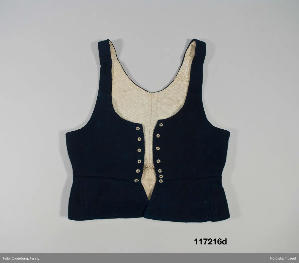 Huvudliggaren:
"Sorgdräkt för kvinna. Särk med 'hängseltåg', 'särkyvdel'; vit -'häsklä'; blått livstycke; svart tröja; huvudduk; 'svart näsduk'; hatt; 'raskförklä', 'sockar',  'hårbandsvifsa', huvudbindel."

Bilaga:
a: "8 Särk, övdel. av linne eller blågarn. med broderade linningar på överdelen"
c: "11. Hvit häsklä."
Anm: Kung Kerstin Larsdotters resa i Floda sn Dalarna 24/2 - 8/3 1912. Innehåller förteckning på inköp av dräktdelar från Floda, inv nr 117 216 - 117 239. Här finns  tillverkningstid och beskrivningar på vissa av dräktdelarna.  I vissa fall nämns också utförligt på vilka helgdagar dräktdelen  användes.

Katalogkort:
a: Hängselsärk
b. Överdel av halvlinne vävt i kypert.
c.  Halskäde av småmönstrat bomullstyg, liknande kambris, vävt  i kypertvariation med drällmönster.
d. Livstycke av mörkblå kraftig vadmal,
e. Tröja av grov svart vadmal

i. 
 Mönstervävt band med upplockat mönster i rött z-tvinnat ullgarn på vit linnebotten, med hjärtan, kryss och rombformer, 2 band om vardera 124 cm har vikts och sytts ihop så att det ena bildar en ögla och det andra har fria ändar avslutade med en tofs i bandets garn.
Läs mer om dräktskicket i Floda i: Albert Alm, Dräktalmanacka för Floda socken, Nordiska museet, Sthlm 1930
/Berit Eldvik 2005-04-14