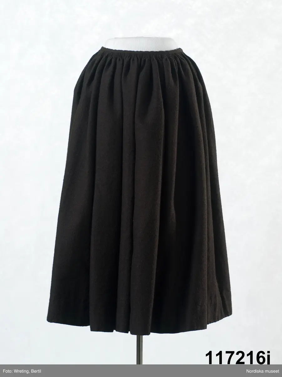 Huvudliggaren:
"Sorgdräkt för kvinna. Särk med 'hängseltåg', 'särkyvdel'; vit -'häsklä'; blått livstycke; svart tröja; huvudduk; 'svart näsduk'; hatt; 'raskförklä', 'sockar',  'hårbandsvifsa', huvudbindel."

Bilaga:
a: "8 Särk, övdel. av linne eller blågarn. med broderade linningar på överdelen"
c: "11. Hvit häsklä."
Anm: Kung Kerstin Larsdotters resa i Floda sn Dalarna 24/2 - 8/3 1912. Innehåller förteckning på inköp av dräktdelar från Floda, inv nr 117 216 - 117 239. Här finns  tillverkningstid och beskrivningar på vissa av dräktdelarna.  I vissa fall nämns också utförligt på vilka helgdagar dräktdelen  användes.

Katalogkort:
a: Hängselsärk
b. Överdel av halvlinne vävt i kypert.
c.  Halskäde av småmönstrat bomullstyg, liknande kambris, vävt  i kypertvariation med drällmönster.
d. Livstycke av mörkblå kraftig vadmal,
e. Tröja av grov svart vadmal

i. 
 Mönstervävt band med upplockat mönster i rött z-tvinnat ullgarn på vit linnebotten, med hjärtan, kryss och rombformer, 2 band om vardera 124 cm har vikts och sytts ihop så att det ena bildar en ögla och det andra har fria ändar avslutade med en tofs i bandets garn.
Läs mer om dräktskicket i Floda i: Albert Alm, Dräktalmanacka för Floda socken, Nordiska museet, Sthlm 1930
/Berit Eldvik 2005-04-14