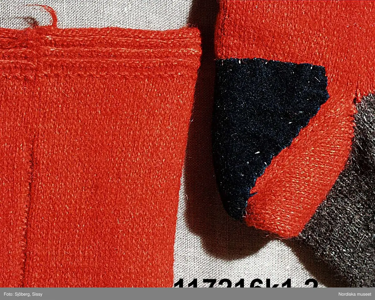 Huvudliggaren:
"Sorgdräkt för kvinna. Särk med 'hängseltåg', 'särkyvdel'; vit -'häsklä'; blått livstycke; svart tröja; huvudduk; 'svart näsduk'; hatt; 'raskförklä', 'sockar',  'hårbandsvifsa', huvudbindel."

Bilaga:
a: "8 Särk, övdel. av linne eller blågarn. med broderade linningar på överdelen"
c: "11. Hvit häsklä."
Anm: Kung Kerstin Larsdotters resa i Floda sn Dalarna 24/2 - 8/3 1912. Innehåller förteckning på inköp av dräktdelar från Floda, inv nr 117 216 - 117 239. Här finns  tillverkningstid och beskrivningar på vissa av dräktdelarna.  I vissa fall nämns också utförligt på vilka helgdagar dräktdelen  användes.

Katalogkort:
a: Hängselsärk
b. Överdel av halvlinne vävt i kypert.
c.  Halskäde av småmönstrat bomullstyg, liknande kambris, vävt  i kypertvariation med drällmönster.
d. Livstycke av mörkblå kraftig vadmal,
e. Tröja av grov svart vadmal

i. 
 Mönstervävt band med upplockat mönster i rött z-tvinnat ullgarn på vit linnebotten, med hjärtan, kryss och rombformer, 2 band om vardera 124 cm har vikts och sytts ihop så att det ena bildar en ögla och det andra har fria ändar avslutade med en tofs i bandets garn.
Läs mer om dräktskicket i Floda i: Albert Alm, Dräktalmanacka för Floda socken, Nordiska museet, Sthlm 1930
/Berit Eldvik 2005-04-14