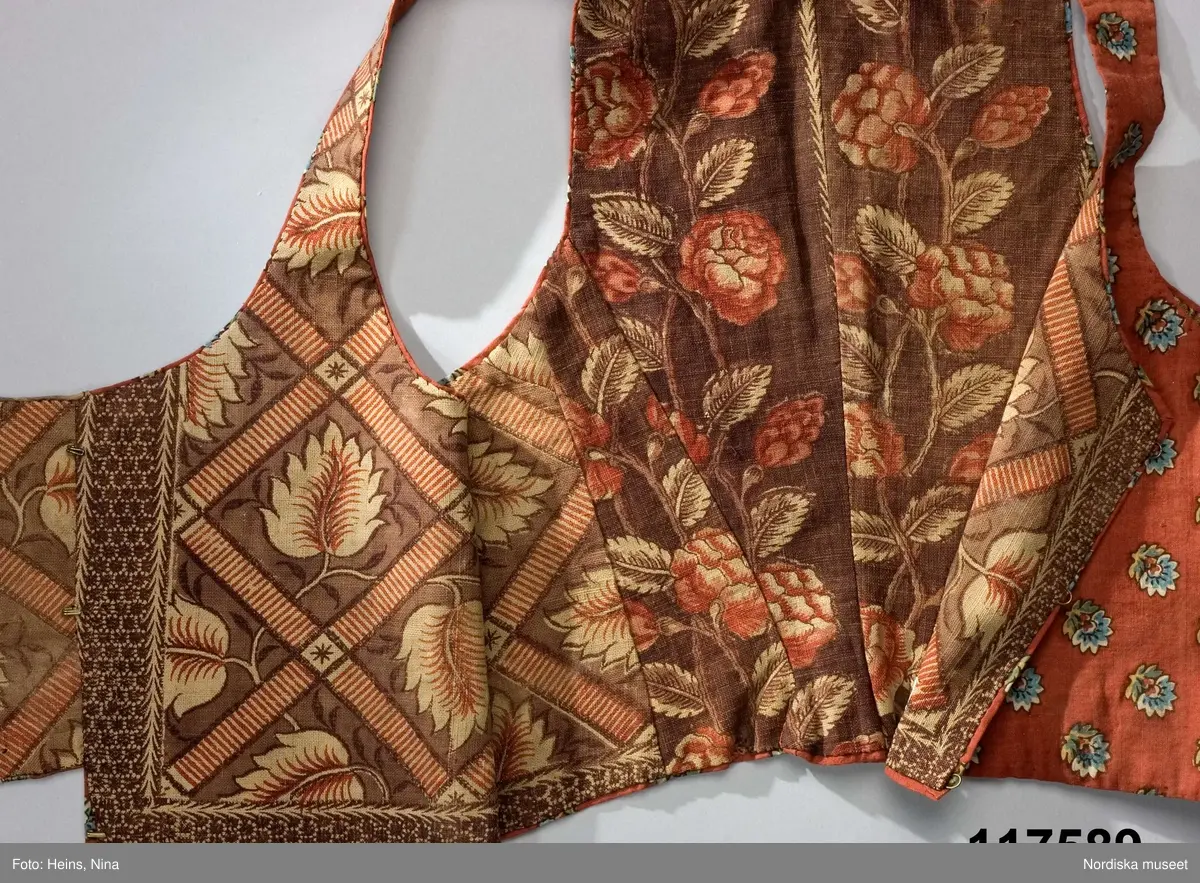 Livstycke av tryckt bomullstyg, kallat kattun, botten färgad i mörk krappröd färg med utsparade små blommor jämnt spridda över ytan. Blommorna har sedan tryckts i med blått och gult med svarta konturer. 2 fram/sidstycken och 4 ryggstycken som avsmalnar ned i ett rudimentärt skört utan veck, små infällda kilar i sidorna. Knäppt med 3 par hakar och hyskor, knäppningen dold under ett förslag av kattuntyget och en bit från ett halskläde i sidensatin med kantbården kvar. Livstycket fodrat med 2 olika kattuntryck av troligen något äldre datum, i ryggen ett brunbottnat bomullstyg med rosenrankor i krapprött och den oblekta grundfärgen, och i framstyckena ett bladmönster i gallerverk i samma färgskala men något ljusare. Troligen är det senare tyget också ett återbrukat halskläde där man skickligt lagt kantbården så att den följer även runt livstyckets kanter.
/Berit Eldvik 2006-12-08