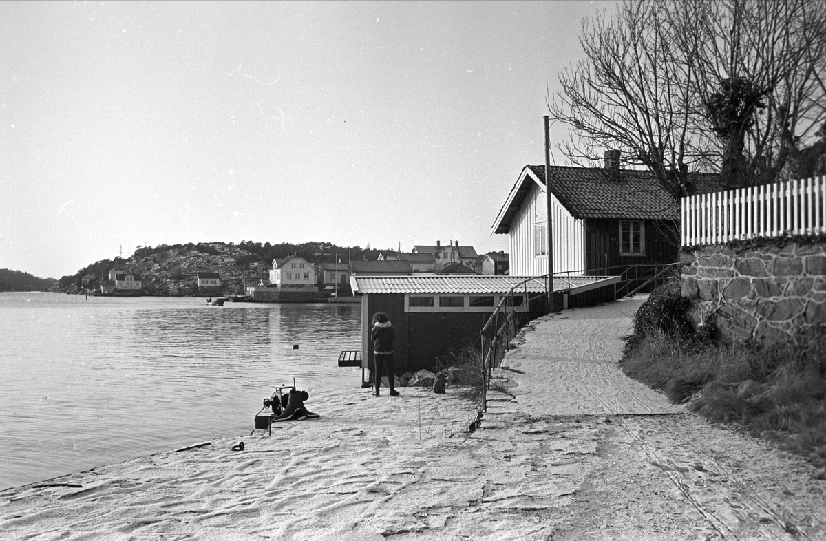 Fra Risør januar 1967. Boliger ved strandkanten, en kvinne står ved et båthus.