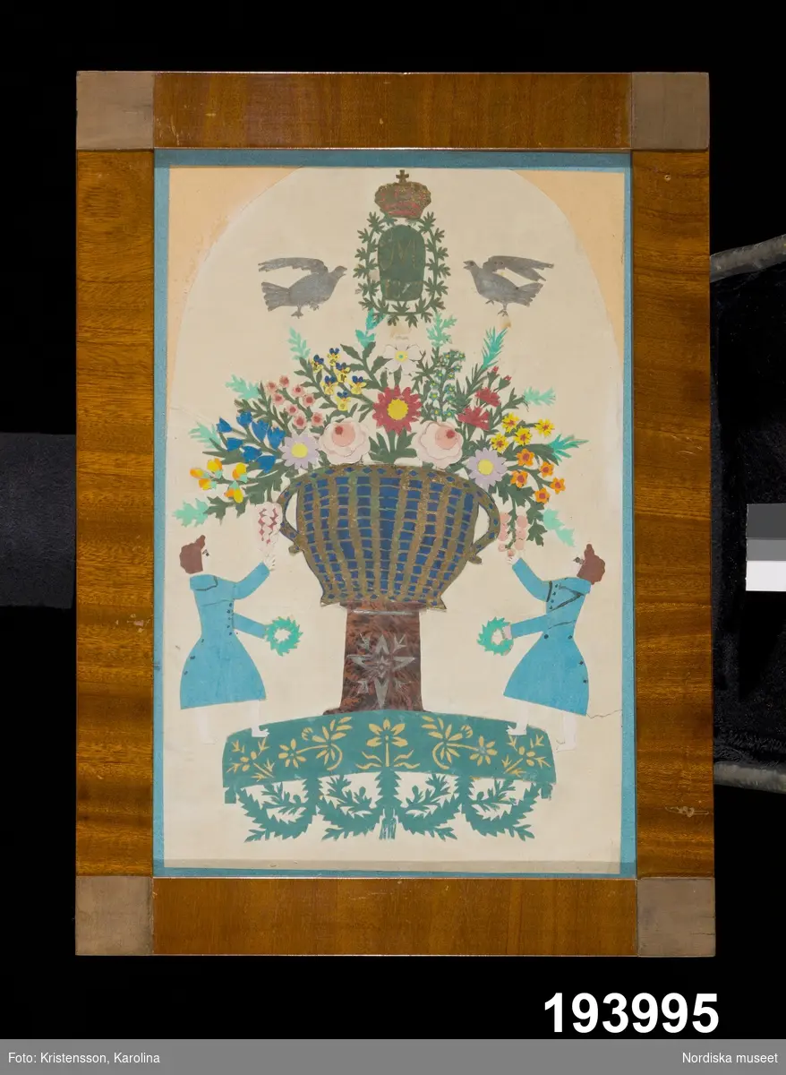 Minnestavla. Blomsterurna på fot flankerad av två män med kransar i händerna.