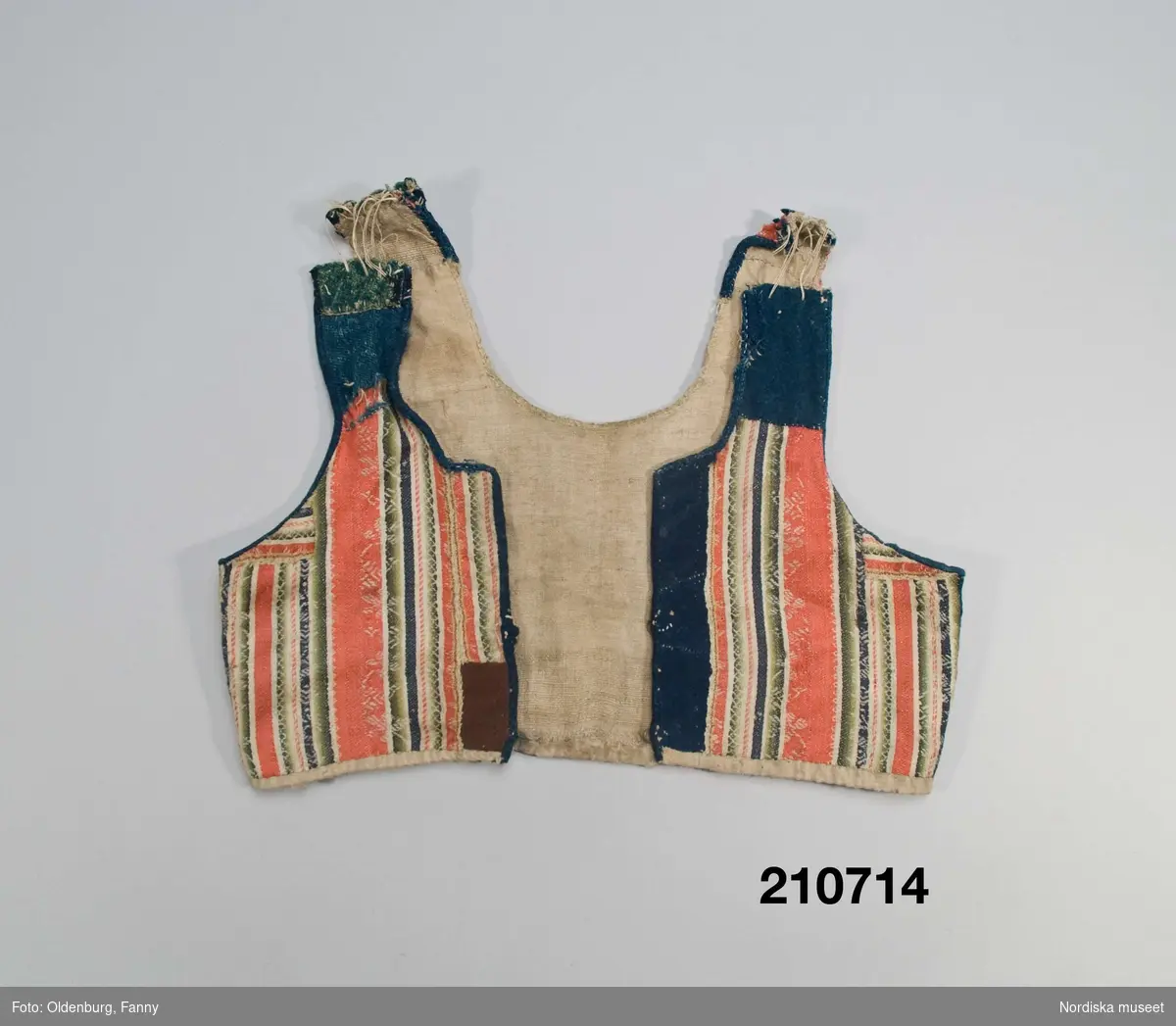 Livstycke "bod" som varit fastsytt på en kjol. Av varprandig yllesatin i rött, blått, vitt och gröna nyanser med invävda vita mönsterbårder, kvaliteten kallas blommerad kalmink och vävdes av kamgarn och glättades. 2 framstycken  och ett bakstycke med 3 påsydda lila band i ryggen. Livstycket består av flera hopskarvade bitar, dels av kalminktyget dels av mörkblått kläde samt blått och grönt vadmal (på axlarna och vid ena framkanten där det även finns en brun bit).I sidorna iskarvad med på ena sidan gult kläde på den andra mörkblått kläde. Kantad kring ärmhål och hals/framkant med blå vadmal och blått kläde. På insidan av framkanterna svart kläde, knäppning med  2 par hakar och hyskor av mässing. Fodrat med oblekt ojämnt vävt linnelärft
Anm. Slitet, avklippt på axlarna där livet förlängts med öglor av dubbelt lingarn.
Sådana blommerade kalminker vävdes främst i England och kom till Norden under 1700-talets andra del och blev moderna i folkligt dräktskick. 
/Berit Eldvik 2007-09-20