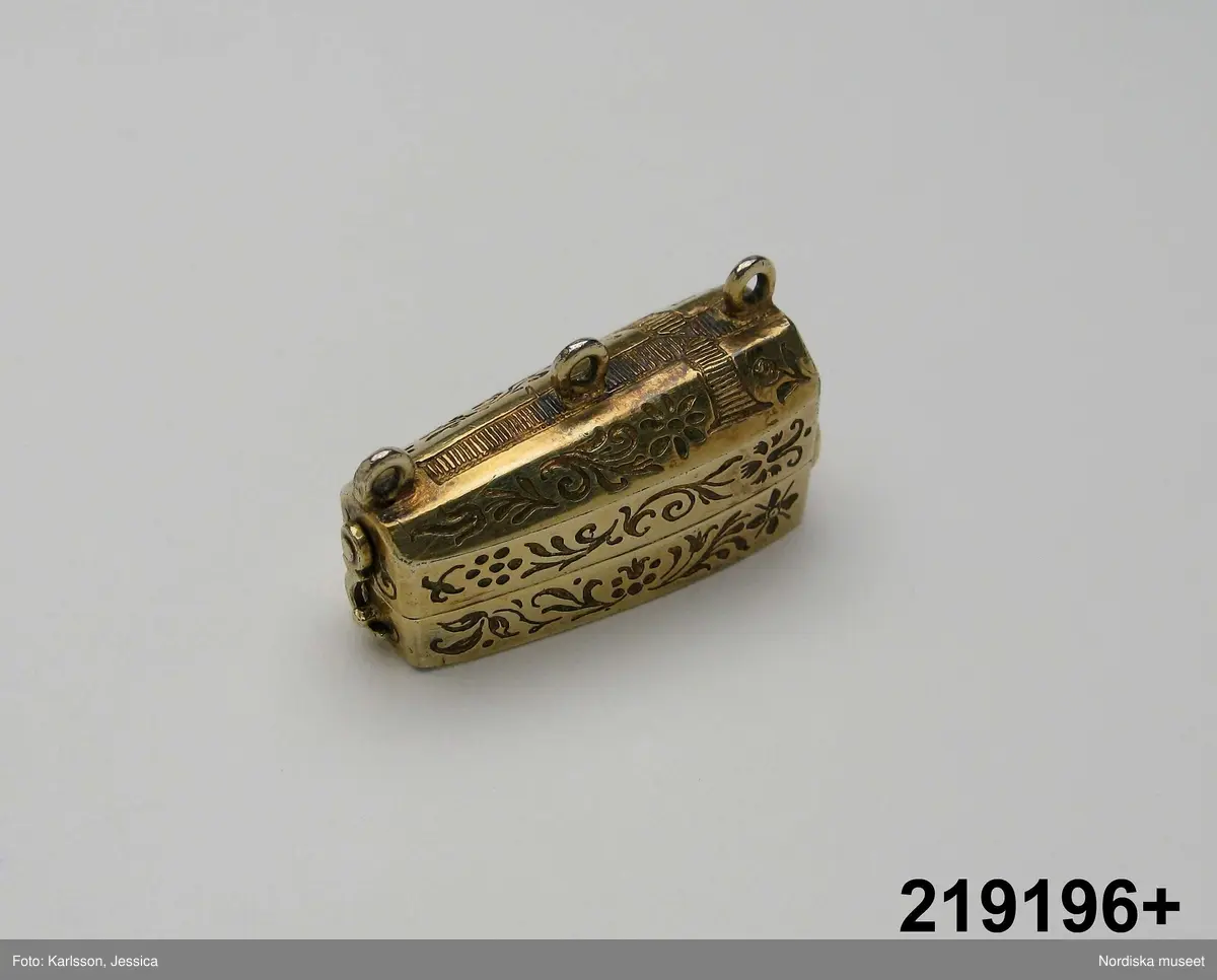 Katalogkort:
"Smycke (berlock)
Silver
L 3,5 cm, br. 1,2 cm
I form av Gustav II Adolfs likkista. Trycklock med haspar. Graverad dekor. Miniatyrbild i trä av kunge, fäst i botten."