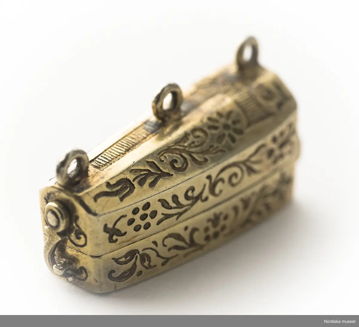 Katalogkort:
"Smycke (berlock)
Silver
L 3,5 cm, br. 1,2 cm
I form av Gustav II Adolfs likkista. Trycklock med haspar. Graverad dekor. Miniatyrbild i trä av kunge, fäst i botten."