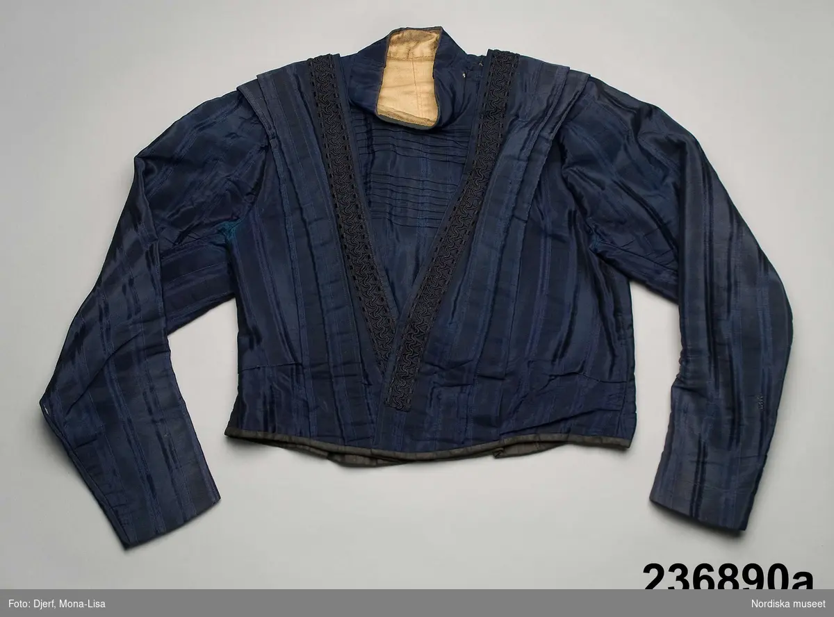 2-delad klänning av mörkblått randvävt halvsiden. a-b samt löst skärp c.

a. Liv figursytt på foderstomme av  ljusgul bomullskypert i fiskbensmönster. 2 framstycken och ett ryggstycke med ett rundat skört bak som dock stoppas ned i kjolen, Fram djupt V-ringad med  2 breda lagda veck som  löper i V-form från axel ned mot underkanten både fram och bak. Längs med vecken även en 3 cm bred  svart maskingjord spets . I ringningen isättning av samma tyg med en mängd tvärgående stråveck, tättslutande ståndkrage  styvad med något mellanlägg. Isydd lång ärm, svängd med 2 sömmar, något rynkad på ärmkullen, snäv vid handleden.
Mitt bak i midjan en fastsydd häkta att fästa mot kjolen. 
b. Kjol av 7 sneddade våder, sydd på foderstomme av svart bomullslärft s.k domestik. Framvåden markerad med nedsydda veck vid sömmarna. Kjolen slät framtill och rynkad bak i midjan mot en 2 cm bred linning av samma tyg. Mitt bak i linningen en fastsydd hyska att häkta mot livet. Dolt sprund i vänster framsöm knäppt med  5 tryckknapapr och överst en hake och hyska. Infälld ficka i sprundet. Skoning i nederkanten av samma tyg som fodret. I kanten slitband med plyschkant.
Anm. 10-12 cm av tyget invikt upptill vid linningen. Kanske ett sätt att inte klippa bort tyg om man skulle vilja återanvända det senare. Det var mycket vanligt att just tygrika kjolar kunde sys om till annat.
c. Löst skärp av samma tyg.  Veckat på längden. Ofodrat, söm mitt bak, 3 fastsydda stålfjädrar på insidan. Knäpps med 3 svarta hakar och tränsade trådöglor 
Klänningen bör vara sydd efter 1903 då Prym uppfann tryckknappen.
/Berit Eldvik 2011-09-08