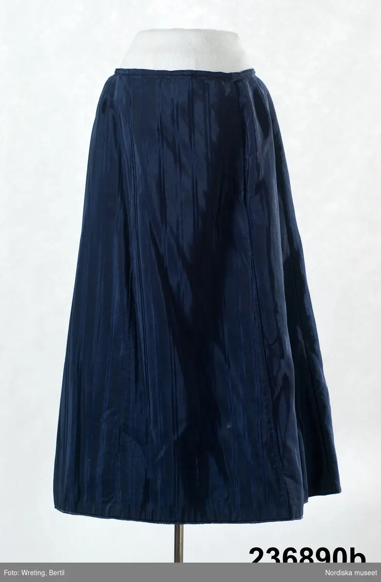 2-delad klänning av mörkblått randvävt halvsiden. a-b samt löst skärp c.

a. Liv figursytt på foderstomme av  ljusgul bomullskypert i fiskbensmönster. 2 framstycken och ett ryggstycke med ett rundat skört bak som dock stoppas ned i kjolen, Fram djupt V-ringad med  2 breda lagda veck som  löper i V-form från axel ned mot underkanten både fram och bak. Längs med vecken även en 3 cm bred  svart maskingjord spets . I ringningen isättning av samma tyg med en mängd tvärgående stråveck, tättslutande ståndkrage  styvad med något mellanlägg. Isydd lång ärm, svängd med 2 sömmar, något rynkad på ärmkullen, snäv vid handleden.
Mitt bak i midjan en fastsydd häkta att fästa mot kjolen. 
b. Kjol av 7 sneddade våder, sydd på foderstomme av svart bomullslärft s.k domestik. Framvåden markerad med nedsydda veck vid sömmarna. Kjolen slät framtill och rynkad bak i midjan mot en 2 cm bred linning av samma tyg. Mitt bak i linningen en fastsydd hyska att häkta mot livet. Dolt sprund i vänster framsöm knäppt med  5 tryckknapapr och överst en hake och hyska. Infälld ficka i sprundet. Skoning i nederkanten av samma tyg som fodret. I kanten slitband med plyschkant.
Anm. 10-12 cm av tyget invikt upptill vid linningen. Kanske ett sätt att inte klippa bort tyg om man skulle vilja återanvända det senare. Det var mycket vanligt att just tygrika kjolar kunde sys om till annat.
c. Löst skärp av samma tyg.  Veckat på längden. Ofodrat, söm mitt bak, 3 fastsydda stålfjädrar på insidan. Knäpps med 3 svarta hakar och tränsade trådöglor 
Klänningen bör vara sydd efter 1903 då Prym uppfann tryckknappen.
/Berit Eldvik 2011-09-08