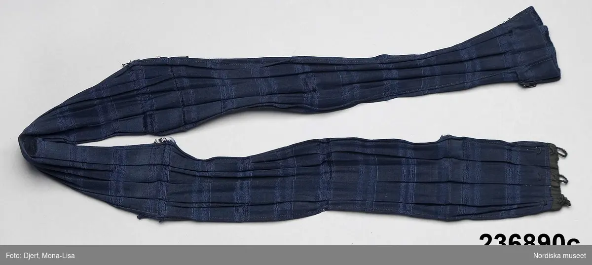 2-delad klänning av mörkblått randvävt halvsiden. a-b samt löst skärp c.

a. Liv figursytt på foderstomme av  ljusgul bomullskypert i fiskbensmönster. 2 framstycken och ett ryggstycke med ett rundat skört bak som dock stoppas ned i kjolen, Fram djupt V-ringad med  2 breda lagda veck som  löper i V-form från axel ned mot underkanten både fram och bak. Längs med vecken även en 3 cm bred  svart maskingjord spets . I ringningen isättning av samma tyg med en mängd tvärgående stråveck, tättslutande ståndkrage  styvad med något mellanlägg. Isydd lång ärm, svängd med 2 sömmar, något rynkad på ärmkullen, snäv vid handleden.
Mitt bak i midjan en fastsydd häkta att fästa mot kjolen. 
b. Kjol av 7 sneddade våder, sydd på foderstomme av svart bomullslärft s.k domestik. Framvåden markerad med nedsydda veck vid sömmarna. Kjolen slät framtill och rynkad bak i midjan mot en 2 cm bred linning av samma tyg. Mitt bak i linningen en fastsydd hyska att häkta mot livet. Dolt sprund i vänster framsöm knäppt med  5 tryckknapapr och överst en hake och hyska. Infälld ficka i sprundet. Skoning i nederkanten av samma tyg som fodret. I kanten slitband med plyschkant.
Anm. 10-12 cm av tyget invikt upptill vid linningen. Kanske ett sätt att inte klippa bort tyg om man skulle vilja återanvända det senare. Det var mycket vanligt att just tygrika kjolar kunde sys om till annat.
c. Löst skärp av samma tyg.  Veckat på längden. Ofodrat, söm mitt bak, 3 fastsydda stålfjädrar på insidan. Knäpps med 3 svarta hakar och tränsade trådöglor 
Klänningen bör vara sydd efter 1903 då Prym uppfann tryckknappen.
/Berit Eldvik 2011-09-08