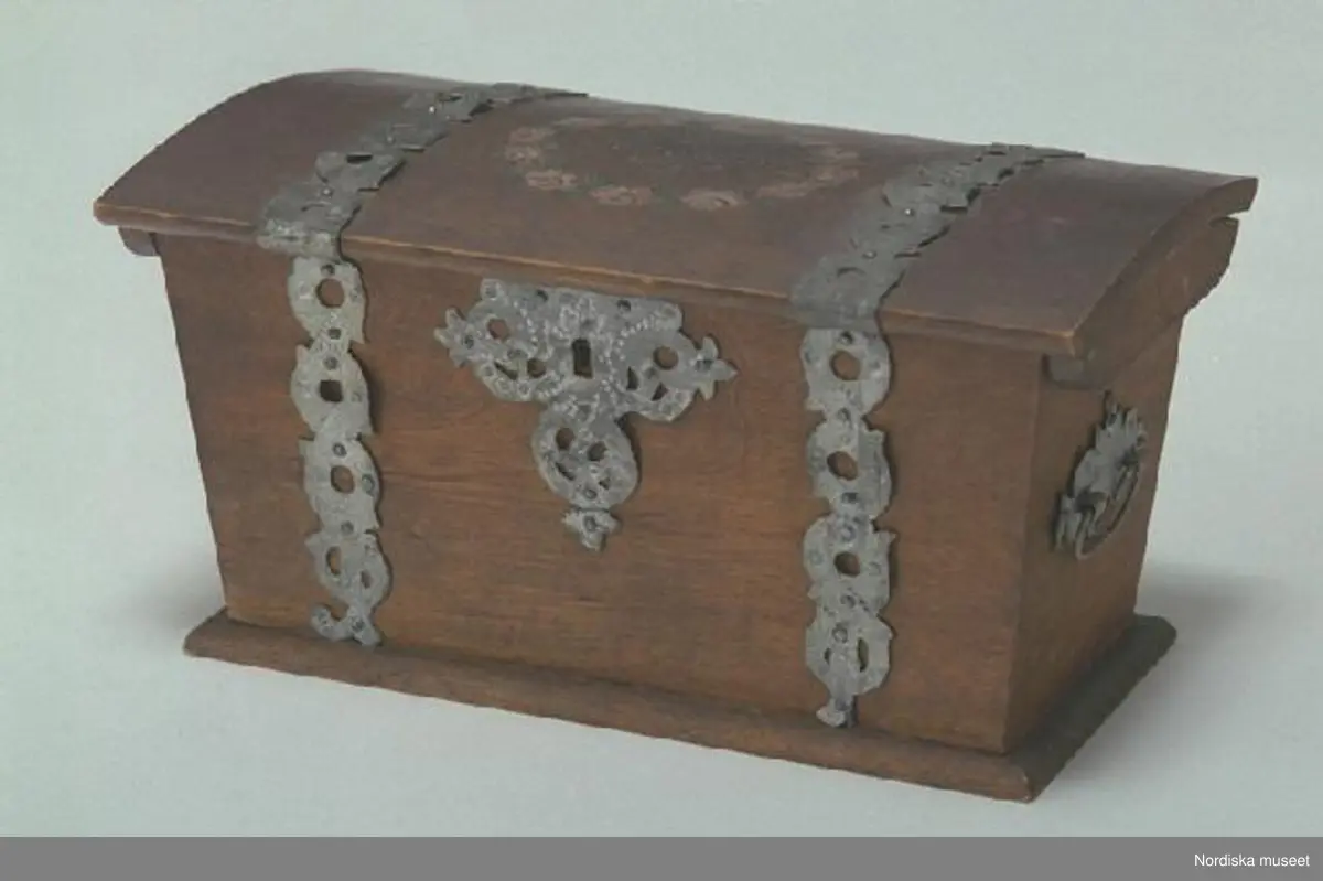 Inventering Sesam 1997-1998:
B 21,5, Djup 12,5, H 11,5 cm
Skrin, rektangulärt, i kistform, med svagt välvt fällock, av lövträ, björk. Utanpåliggande botten, fästad med lim, överskjutande kanter nedtill som fotlist. Sidorna ihopsatta med lim.
Locket har gavlar limmade på undersidan, två gångjärn av mässing fästade invändigt med skruvar. Instickslås av mässing, med låssprint av mässing i locket, lås och sprint fästade med skruvar, nyckel saknas.
Utvändigt prytt på lock och sidor med utstansade beslag av järn, silverfärgad yta, nyckelskylt och bygelhandtag av på gavlarna.
Utsidan brunbetsad, möjligen för att imitera ek. På locket en blomsterkrans, rosor i vitt och rosa med gröna blad, inuti kransen målat med vitt och svart " Anno 1909 ".
Invändigt vid ena gaveln har funnits en läddika, nu sitter endast fällocket kvar medan de två andra bitarna ligger lösa i skrinet, har varit limmade.
I skrinet har förvarats lös tobak, i läddikan cigarettpapper.
Från Bromanska sängkammaren.
Agnetha Blomberg 1998