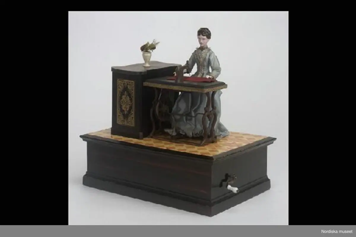 Inventering Sesam 1996-1999:
L  24,8   B  17,7   H  25 (cm)
Speldosa (a), leksak, damdocka sittande vid symaskin på rektangulär, vevf¿rsedd låda av brunbetsat och lackat barrträ.
Docka av målad papier mach¿ med brunt uppsatt hår, ljusblå sidenklänning med turnyr, trampsymaskin av brunmålat trä; gråmålat bord med konturerat underrede, trampa och vevhjul, symaskinsarm med nål, r¿tt tygstycke, träpall på tre ben med r¿d sammetssits, skåp av brunbetsat och lackat trä med dekor i guldfärgat ¿verf¿ringstryck, kruka av trä med blomma av papper, textil och fjäder, imiterad korkmatta av lackat papper med tryckt geometriskt m¿nster i gult och orange. 
F¿rvaringslåda med skjutlock (b+) av obehandlat barrträ, tryckt etikett med motiv av leksaker samt diverse påskrifter, bl a "1/12 dz 1782".
Vid gång r¿r sig fottrampan, nålen går upp och ned och tyget f¿rflyttar sig.
Ägarinnan fick speldosan i gåva av sina f¿räldrar när hon var barn på 1890-talet, då familjen bodde i Kalmar. M¿jligen tysk tillverkning.
Anna Womack feb 1998