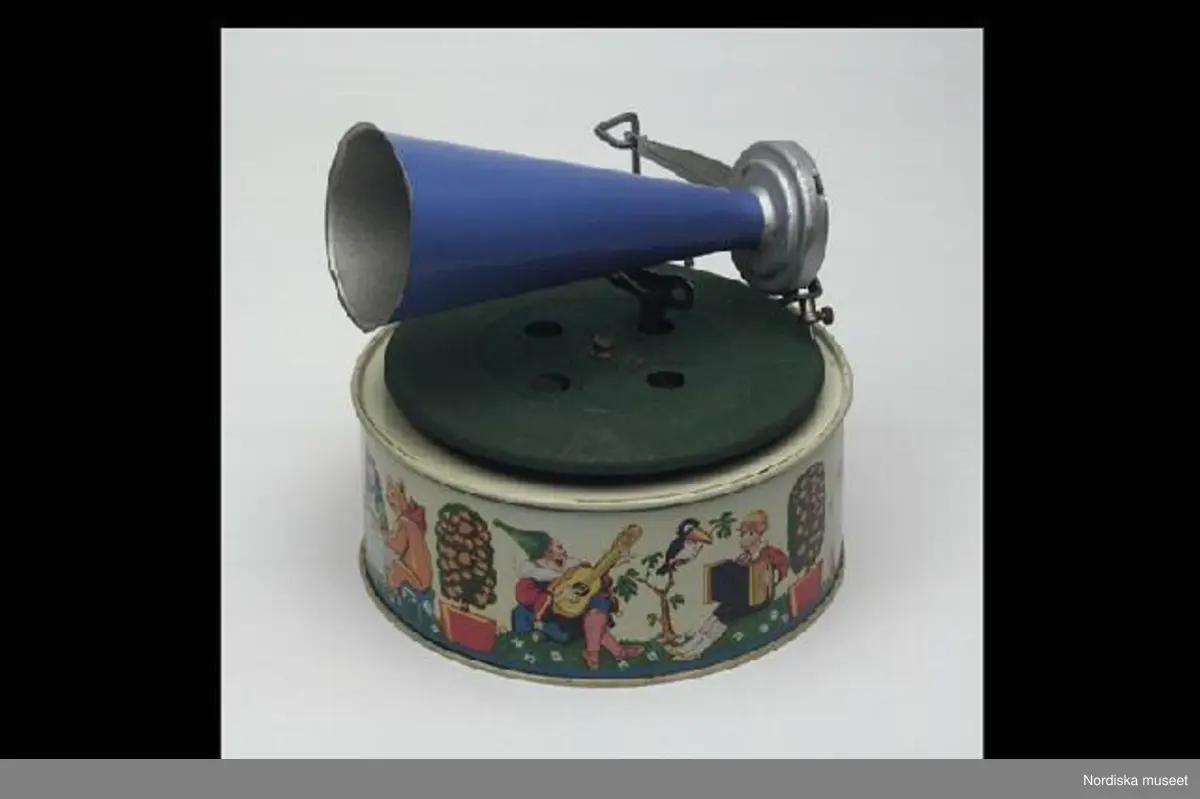 Inventering Sesam 1996-1999:
Diam  16,5   H  17 (cm)
Trattgrammofon, leksak, av bockad och lackerad plåt, rund ljuddosa med bilder av lekande och musicerande barn, djur och clowner i litografiskt tryck mot ljusgul fond, ovanpå denna en roterande platta, täckt med grönt textilstoff, rörlig arm med nål och blå tratt, stoppspak på ljuddosan, lös nyckel. Märkt: "Keimola / D.R.G.M. Registered. / Made in Germany".
Har tillhört givarinnans kusin, elektrikern Tage Fransson (1918-1971), som barn. Dennes far var skräddare och bodde på Västgötagatan, Stockholm.
I-A Berg [=Inga Arnö-Berg]
Anna Womack feb 1998