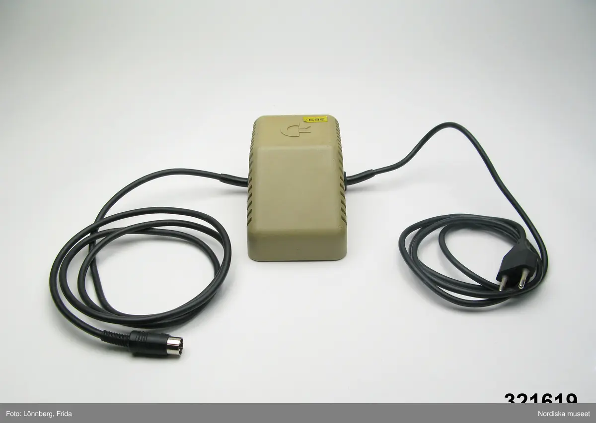 Inventering Sesam 1996-1999:
L 13,5 cm
B 8,5 cm
H 7 cm
Transformator med grått plasthöljje. Randig på sidorna, Commodores logotype ovanpå; ett C med två streck (C=, ungefär). Baktill svart avstängningsknapp och text "TRANSFORMATOR / PASRT - NO 902503-11" mm. F¿r 5 volt - 1,5 ampère. Två fasta svarta sladdar, en med stickkontakt och en med DIN-kontakt. Ovantill gul pappersetikett "369". Undertill stämplad i gult "20 (eller 26) APRIL 1985".
Axel Brundin (född 1977) fick datorn i julklapp. Den var köpt från pojkarna Norman (födda 1971) som tidigare använt den.
Dataspel och tillhörande föremål; Tangentbord ivn.nr 321.617 kasettbandspelare inv.nr 321.618, transformator inv.nr 321.619, Kasettväska med kasettband med olika dataspel inv.nr 321.620:1-24, tre joy sticks inv.nr 321.621 och 321.622, skokartong inv.nr 321.623 (i vilken kasettband f¿rvarades)  och Kasettband med spel 321.624-321.639
Instruktionsböcker, band- och kundförteckning finns på arkivet.
Bilaga
EB 1995  [=Elisabeth Brundin]
Leif Wallin 1998