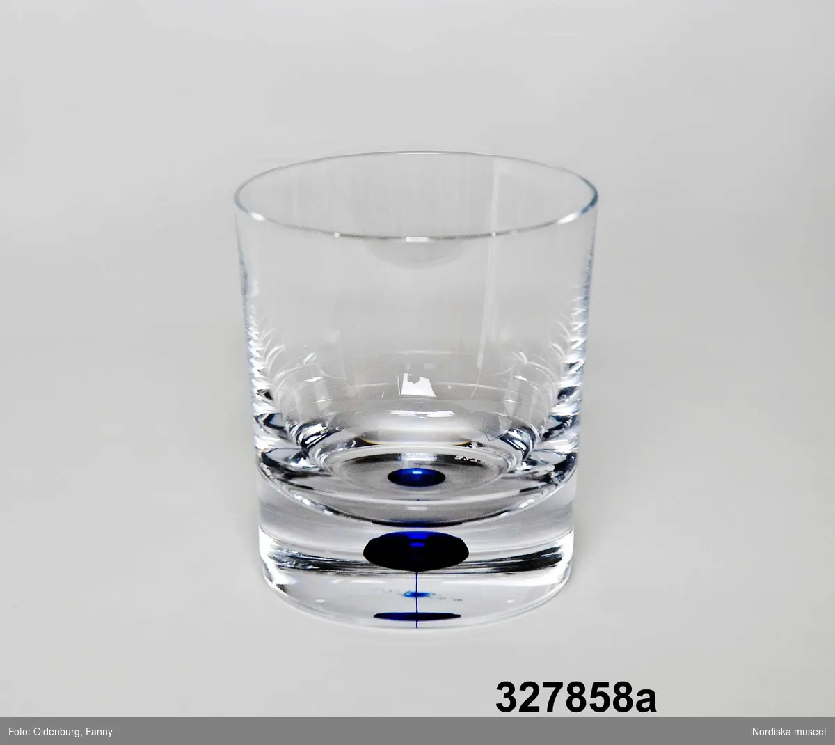 Huvudliggaren:
"Dricksglas i ask, 2 st, glas, papp. Klart, cylindriskt, koboltblå bubbla i tjock botten. Form Erik Lagerbielke Intermezzo, tillv 2000 Orrefors, etsad tillvsign. Vinröd ask, viklock. Folder. Anv 2001 under Sveriges EU-ordförandeskap. Old-Fashioned."

Kataloglapp:
Old fashioned-glas med tjock botten, koboltblå bubbla i botten.
Form Erika Lagerbielke, 1985 "Intermezzo"
Vinröd originalkartong.

Ingick i den kuvertuppsättning som utformades av Sveriges Lantbrukares Riksförbund (LRF) och användes vid ett antal av de officiella middagar som anordnades av svenska staten under Sveriges ordförandeskap i Europeiska unionen.. 
Se även inv nr

Ingår i ett större förvärv av utrustning och möbler som användes vid den officiella representationen i samband med Sveriges ordförandeskap i Europeiska unionen, våren 2001.
Se även inv nr

Se även Nordiska museets arkiv 6/2002 för informations- och dokumentationsmaterial.
/Helena Ulander/Cecilia Hammarlund-Larsson 2002
Förpackning: Munksjö förpackningar
/Heidi Henriksson/Kersti Wikström 2004
