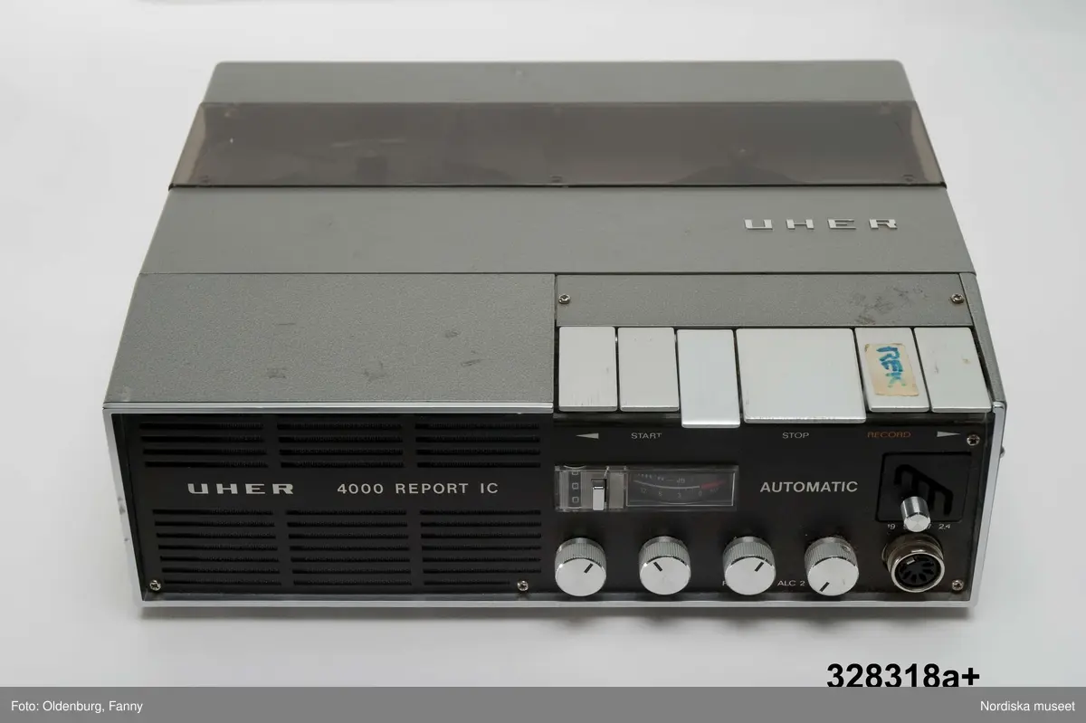 a-g:
Bärbar bandspelare för rullband. Metall, rektangulär, grå eloxerad, tangenter, vred på långsidan, fönster med ljudskala, varvräknare. Spole, mikrofon, mikrofonstativ, bruksanvisning. Batteri- och nätdriven, laddningsbar.
Tillhörande brun läderväska, axelväska med bred rem, ficka för bruksanvisning, mikrofonfack, konstmaterialfoder, uppknäppbar framsida, fällock med spännen. Adresslapp handskriven med förste intendet Göran Rosanders handstil.

a+) Bandspelare
b) Batteriladdare med sladd
c) Mikrofon med sladd
d) Mikrofonstöd
e) Intruktionsbok
f) Kopplingsschema (papper)
g+) Väska.

Den bärbara Uher-bandspelaren innebar en revolution för fältarbete. Fältarbetaren var inte enbart hänvisad till papper och penna. På 1970-talet fanns till en början endast en bärbar bandspelare. Fältarbeten planerades efter tillgänglighet till bandspelare. Samtliga avdelningar samsades till en början om dennna.
/Heidi Henriksson 2005-10-11