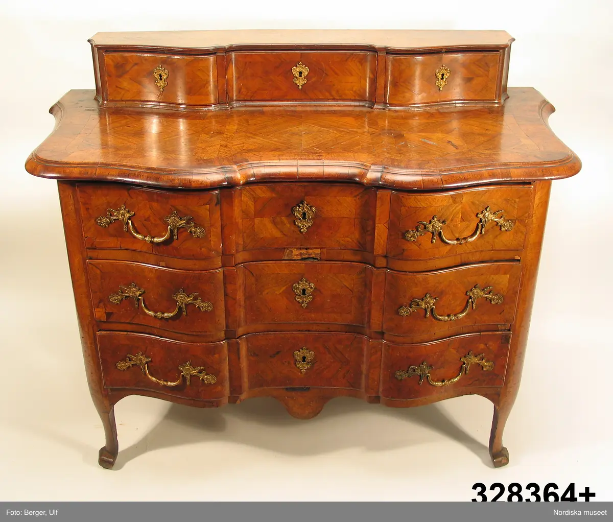 Trelådig byrå, senbarock/Fredrik I:s tid, 1730-40-tal, enkelsvept, med arbalète-formad svängd front. Uppsats med tre lådor. Fanerad på furu med valnöt. Faneret på lådfronterna lagt i kvadrerade fält med fris. Svängda ben med klubbfötter. Utdragbar skrivskiva fanerad med valnöt på ovansidan. Franska régencebeslag av gjuten mässing. Lådbottnar och lådsidor av al. Hela fackbottnar.

Brännstämplad tre gånger på ryggen: "I.H.F" (Johan Hugo Fürloh, mästare i Stockholms snickareämbete 1724-1745. Verkstaden drevs vidare av änkan till 1747).

Anm: Mellersta lådans baksida och botten maskäten. På ryggen etikett med påskrift: "Cyanvätesgasbehandlad  den 26/2 1938 / Hälsovårdsnämndens Cyanvätesanstalter".
Två bygelhandtag nytillverkade juni 2005 på B A konstgjuterier AB, Rosenlundsgatan, Stockholm (märkta med inv.nr och ' Nytillv. 2005' ).  
/Anna Womack 2006-03-02