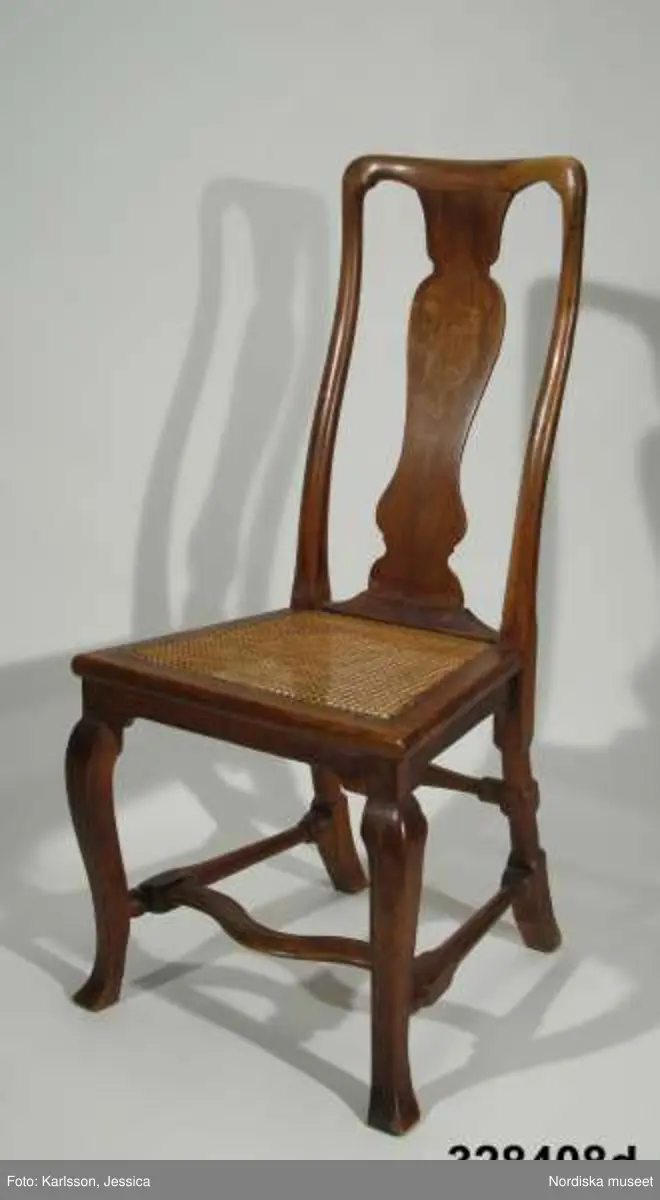 Stol, 6 st (a-f), England, 'Queen Anne', omkring 1720, av valnöt. Hög, balusterformad ryggbricka med inlagd (inkrusterad) dekor av ljusare träslag. Cabriolformade ben och svängt fotkryss med svarvade sidoslåar och bakre tvärslå. Sits med låg sarg, rottingflätad (rottingflätningen nygjord omkring 2002). Tillverkarinitialer inslagna på ben, ryggståndare och baksarg:
a) 'SH' (alt 'HS' ) samt 'IB'.
b) Rund stämpel: [kryss med dubbla streck] 
c) 'SH' (alt 'HS) samt 'IB'
d) 'SH' (alt HS') samt 'IB'
e) 'RH' (3 gånger) 
f) 'RH' (3 gånger),  'IB' (2 gånger) samt 'SH' (alt 'HS')

Jämför stol, inv nr 104.416 med initialerna 'RH' (enligt katalogkort). De engelska stolarnas märkning med tillverkarinitialer på t ex ben, sarg och ryggståndare visar på en rationalisering i tillverkningsprocessen. Initialerna är ibland placerade uppochnedvända vilket indikerar att de slogs in innan stolen sattes ihop. (se 'English furniture 1660-1714 - From Charles II to Queen Anne', Adam Bowett, Italien 2002, s. 264).
Anm: Flera stolar kompletterarade med delar av nytt trä, ilusade bitar. e) bakre tvärslå sekundärt förstärkt med skruv.
/Anna Womack 2008-09-25

Nordiska museet erbjöds 1992 att som testamentarisk gåva efter då framlidna Louise de Kiriline, Ontario, Kanada, ta emot en grupp sittmöbler bestående av två karmstolar och sex stolar. I samtal mellan arvinge till testator och dåvarande avdelningschefen Ingrid Bergman framgick att bakgrunden till gåvan var att del/delar av möblemanget redan skulle finnas i museets samlingar. En från Kanada översänd polaroidbild visade att en eller möjligtvis två stolar av denna typ, troligen tilllverkade i England omkr. 1720, förvärvats av museet redan 1905. Eftersom möbelgruppen skulle ge ett välkommet tillskott av engelska möbler till Charles Totties malmgård på Skansen tackade Nordiska museet ja till den erbjudna gåvan. Stolarna, som vid ankomsten till Sverige visade sig ha sekundära sitsar, har genom konservatorn Sören Lilja vid Skara museum åter försetts med rottingflätade sitsar av ursprunglig typ. Enligt testators anteckningar hade möblerna en gång tillhört den berömda romanförfattarinnan Sophie von Knorring (1797-1848). På sitt fäderne tillhörde hon ätten Zelow, som under 1700-talet haft stark förankring i Västergötland och även på hennes mans sida var det västgötska inslaget påfallande. De båda stolarna som redan tidigare tillhörde Nordiska museet, inv nr 104.415-104.416, inköptes 1905 från Göteborgs Historiska Museum dit de kommit från den kände samlaren Magister Nilsson i Skara. Dessa stolar är av samma typ, men olikheter i detaljer visar dock att de sannolikt ej ingått i samma möblemang. "Magister Nilsson", adjunkt vid Skara läroverk, som var en eldsjäl i Hazelius anda samlade kulturföremål från sin västgötska hembygd och de stora överensstämmelserna mellan dessa stolar och de stolar som nu förvärvas, visar att typen förekommit i Sverige under 1700-talet och kanske särskilt i de västra landskapen kring västerhavsporten Göteborg. Förvärvet behöver därför inte ses enbart relaterat till Tottieska malmgårdens möblering./Per Falck och Birgitta Martinius 2002.

Testator Louise de Kiriline Lawrence (född Flach, 1894-1992), var född i Sverige och växte upp på godset Svensksund på Vikbolandet i Östergötland. Efter att ha arbetat som röda korssyster under 1:a världskriget och i Ryssland efter revolutionen emigrerade hon till Kanada 1927. 