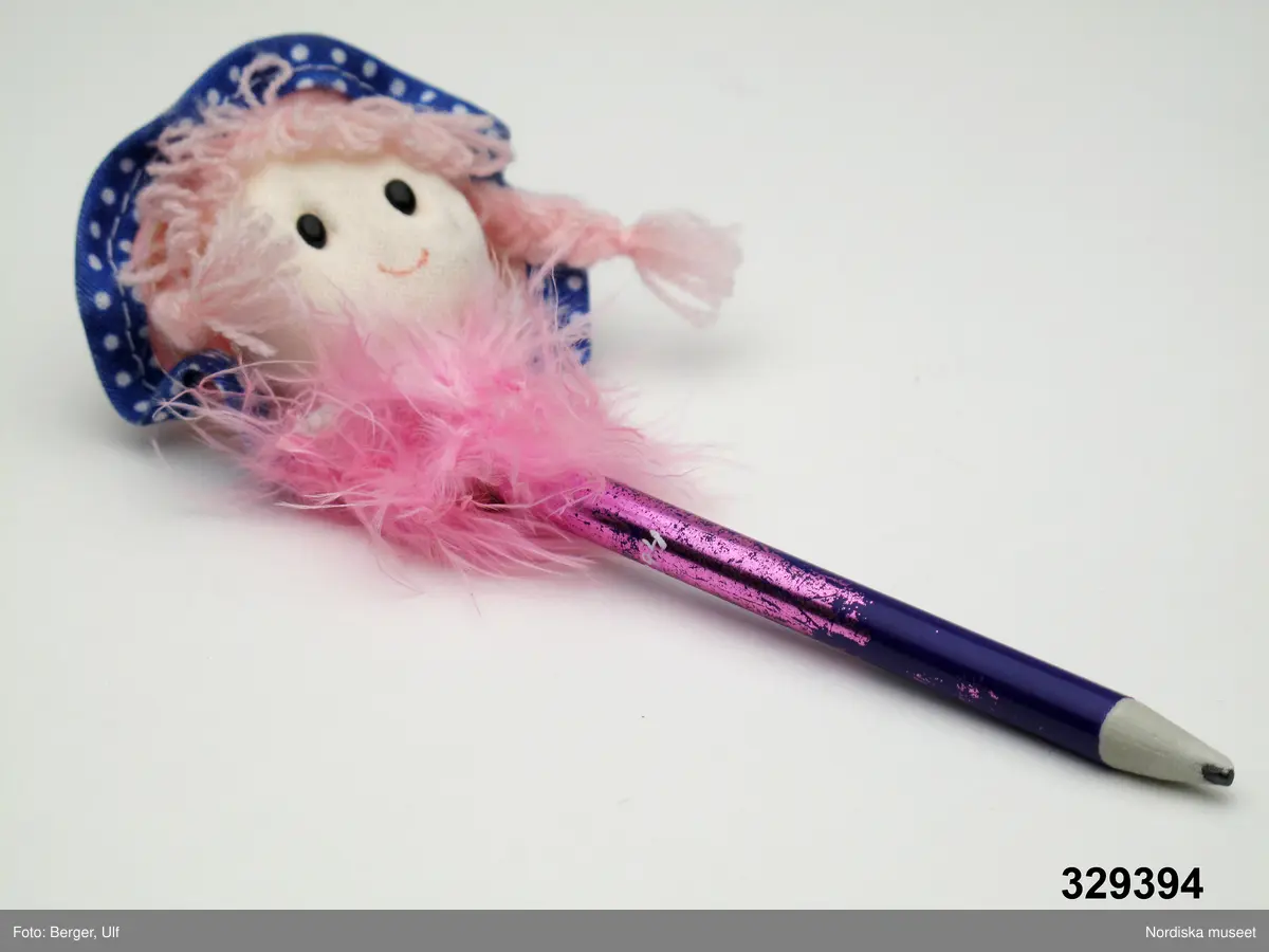Blyertspenna, lila, av konstmaterial, krönt av ett dockhuvud av frigolit täckt med textil. Huvudet med blåbrämad rosa hatt och rosa hår med flätor. Lila ögon och markerad glad mun. Krage av rosa dun mellan penna och huvud.

Givaren (född 1996) upplyser att hon fick föremålet av sin mormor och blev mycket glad för pennan. Tyvärr var dockan inte så bra att skriva med, trots att den vickade fint på huvudet när hon skrev. Men eftersom pennan ändå är så fin så passar det ju perfekt att den bevaras i Nordiska museets samlingar.

Föremålet ingår i projektet "Barn tar plats". Se dokumentation i arkivet D455.
/Ulf Hamilton 2008-05-23