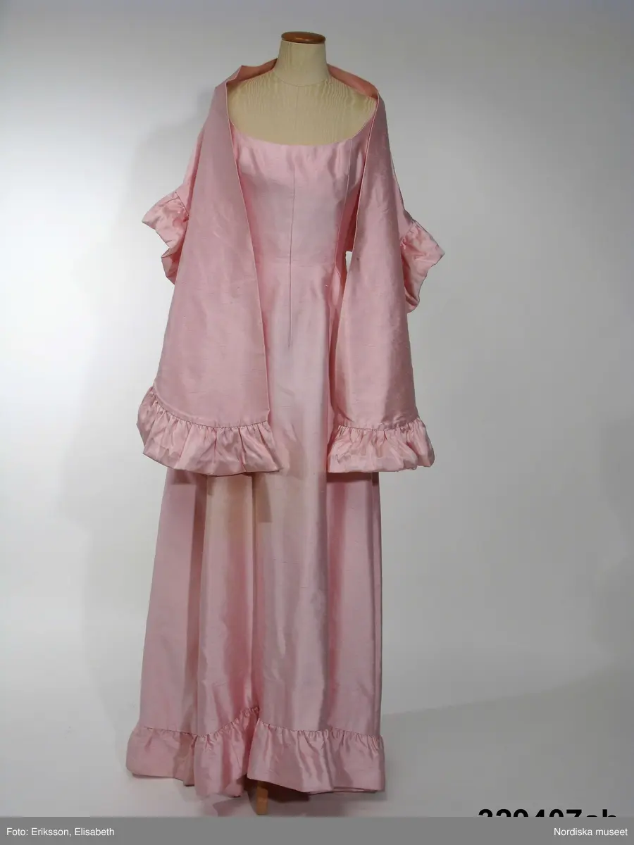 Aftonklänning med Sjal.
a) Aftonklänning av blekt rosa shantungsiden. hellång modell med baddräktsringat åtsittande liv och vid kjol. Helskuren med vid, båtformig urringning som är djupare bak och löper i ett med smala axelband. Bystinsnitt samt längsgående insyningar över byst , mitt fram samt på ryggstycket. Framflyttade sidsömmar. Omlottskuren baktill med mjukt rundad bakvåd. Efter denna från midjan och ner runt kjolen löper en volang av dubbelt tyg. Blixtlås i vänster sida.
Klolen fodrad med styvt nylonnät. Infodringar efter livets urringning, nedtill på kjolen och efter hela omlottvåden. Ryggstycket dubblerat på ett intrikat sätt. 
b) Schal av samma tyg som klänningen, långsmal modell med ett rundat hörn i vardera änden och med volang efter ena långsidan.
Bars med specialkomponerat smycke av glaspärlor.
Komponerad av Gunilla Pontén till Karin Hyldgaard-Jensen, vinnare av Fröken Sverige-titeln 1962. Vinnaren tävlade senare under året om titeln "Miss International Beauty" i Long Beach, Californien. Där kom Karin med till semifinalen. Hela resegarderoben komponerades av Gunilla Pontén. Den kompletterades med konfektionsplagg som också valdes ut av G.P. Till detta kom också en "folkdräkt" som var tung och varm och inte särskilt populär hos Karin. 
 Se inv.nr 329.405-329.407 samt tillbehör 329.585-329.587
/Ingrid Roos2009-06-23