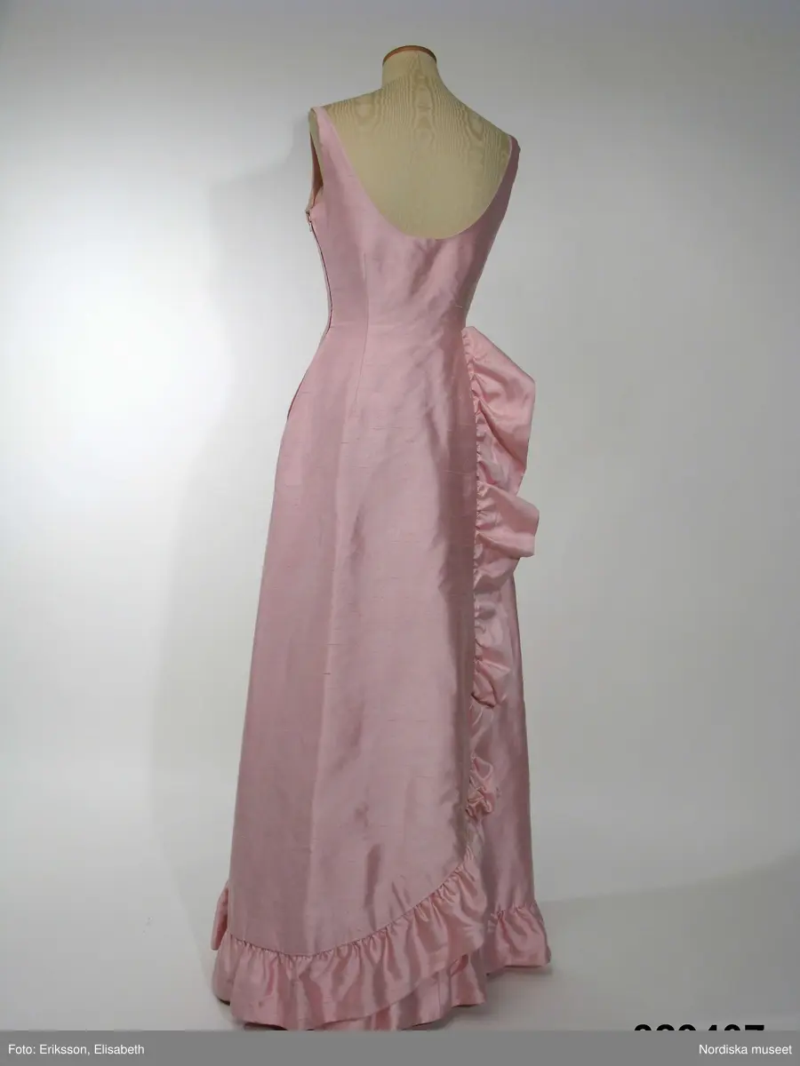 Aftonklänning med Sjal.
a) Aftonklänning av blekt rosa shantungsiden. hellång modell med baddräktsringat åtsittande liv och vid kjol. Helskuren med vid, båtformig urringning som är djupare bak och löper i ett med smala axelband. Bystinsnitt samt längsgående insyningar över byst , mitt fram samt på ryggstycket. Framflyttade sidsömmar. Omlottskuren baktill med mjukt rundad bakvåd. Efter denna från midjan och ner runt kjolen löper en volang av dubbelt tyg. Blixtlås i vänster sida.
Klolen fodrad med styvt nylonnät. Infodringar efter livets urringning, nedtill på kjolen och efter hela omlottvåden. Ryggstycket dubblerat på ett intrikat sätt. 
b) Schal av samma tyg som klänningen, långsmal modell med ett rundat hörn i vardera änden och med volang efter ena långsidan.
Bars med specialkomponerat smycke av glaspärlor.
Komponerad av Gunilla Pontén till Karin Hyldgaard-Jensen, vinnare av Fröken Sverige-titeln 1962. Vinnaren tävlade senare under året om titeln "Miss International Beauty" i Long Beach, Californien. Där kom Karin med till semifinalen. Hela resegarderoben komponerades av Gunilla Pontén. Den kompletterades med konfektionsplagg som också valdes ut av G.P. Till detta kom också en "folkdräkt" som var tung och varm och inte särskilt populär hos Karin. 
 Se inv.nr 329.405-329.407 samt tillbehör 329.585-329.587
/Ingrid Roos2009-06-23