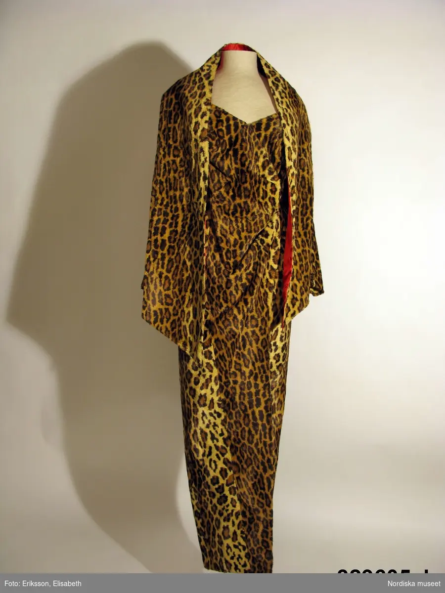 Klänning med tillhörande sjal.
a) Hellång klänning av leopardmönstrad syntetplysch. Figurformad smal modell med lång ärm och öppen omlottslits mitt fram. Främre delningssömmar med bystinsnitt, sidsömmar, söm mitt bak med isytt blixtlås, vertikala bakre insyningar. Bred urringning omlottgående fram med rynkat parti i midjehöjd. Isydd ensömsärm med vid kulle som rynkats mot axelpartiet. Blixtlås i sömmen vid ärmens nederkant. Klänningens liv/kjoldel fodrad med rött fodersiden av viskos, s.k. bembergsiden. 
b) Tillhörande lös schal av samma tyg med rött foder. Rektangulär modell med sneddad nederkant och något smalare mitt bak.

Sydd hos Thalia/Göran Alfredsson 1979 och använd av Alice Timander, tandläkare och känd besökare av de flesta premiärevenemang. Klänningen sydd till premiären på musikalen "Animalen" 1979 på Stora Teatern i Göteborg. Använd även senare.
/Ingrid Roos 2009-09-17.
