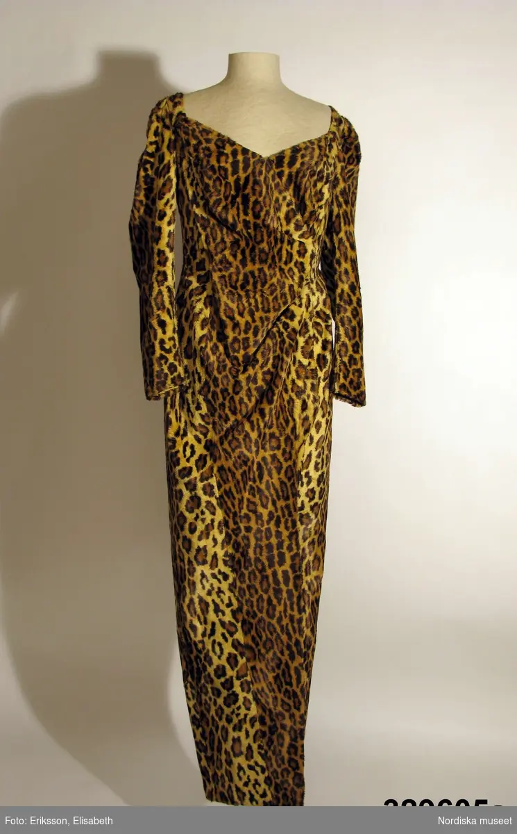 Klänning med tillhörande sjal.
a) Hellång klänning av leopardmönstrad syntetplysch. Figurformad smal modell med lång ärm och öppen omlottslits mitt fram. Främre delningssömmar med bystinsnitt, sidsömmar, söm mitt bak med isytt blixtlås, vertikala bakre insyningar. Bred urringning omlottgående fram med rynkat parti i midjehöjd. Isydd ensömsärm med vid kulle som rynkats mot axelpartiet. Blixtlås i sömmen vid ärmens nederkant. Klänningens liv/kjoldel fodrad med rött fodersiden av viskos, s.k. bembergsiden. 
b) Tillhörande lös schal av samma tyg med rött foder. Rektangulär modell med sneddad nederkant och något smalare mitt bak.

Sydd hos Thalia/Göran Alfredsson 1979 och använd av Alice Timander, tandläkare och känd besökare av de flesta premiärevenemang. Klänningen sydd till premiären på musikalen "Animalen" 1979 på Stora Teatern i Göteborg. Använd även senare.
/Ingrid Roos 2009-09-17.
