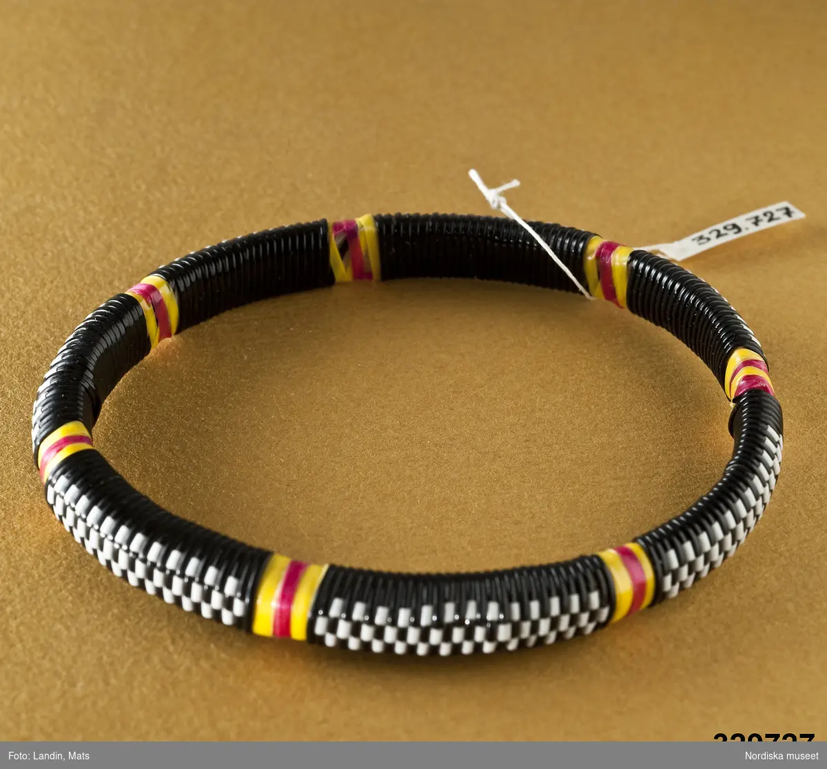 Runt armband med mönster vävt av tunna plastband. Mönster i form av svart/vita rutor och röd/gula band. Afrikanska influenser. Lika som inv.nr 329.726.
/Zingoalla Rosenqvist 2009-02-05